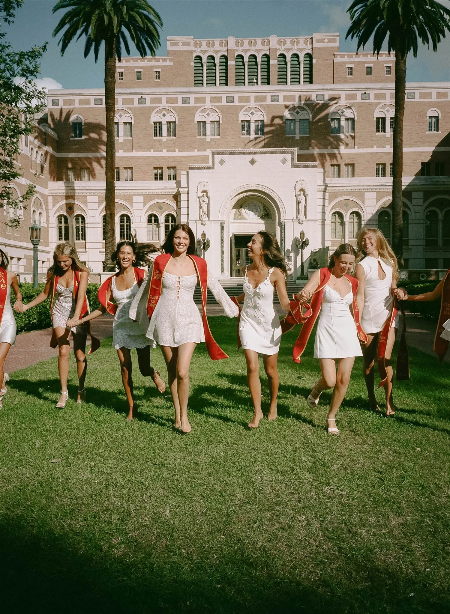 A group of young women in white dresses and red sashes walking in a circle on a green lawn in front of a large historic building with palm trees, arched windows, and decorative statues.