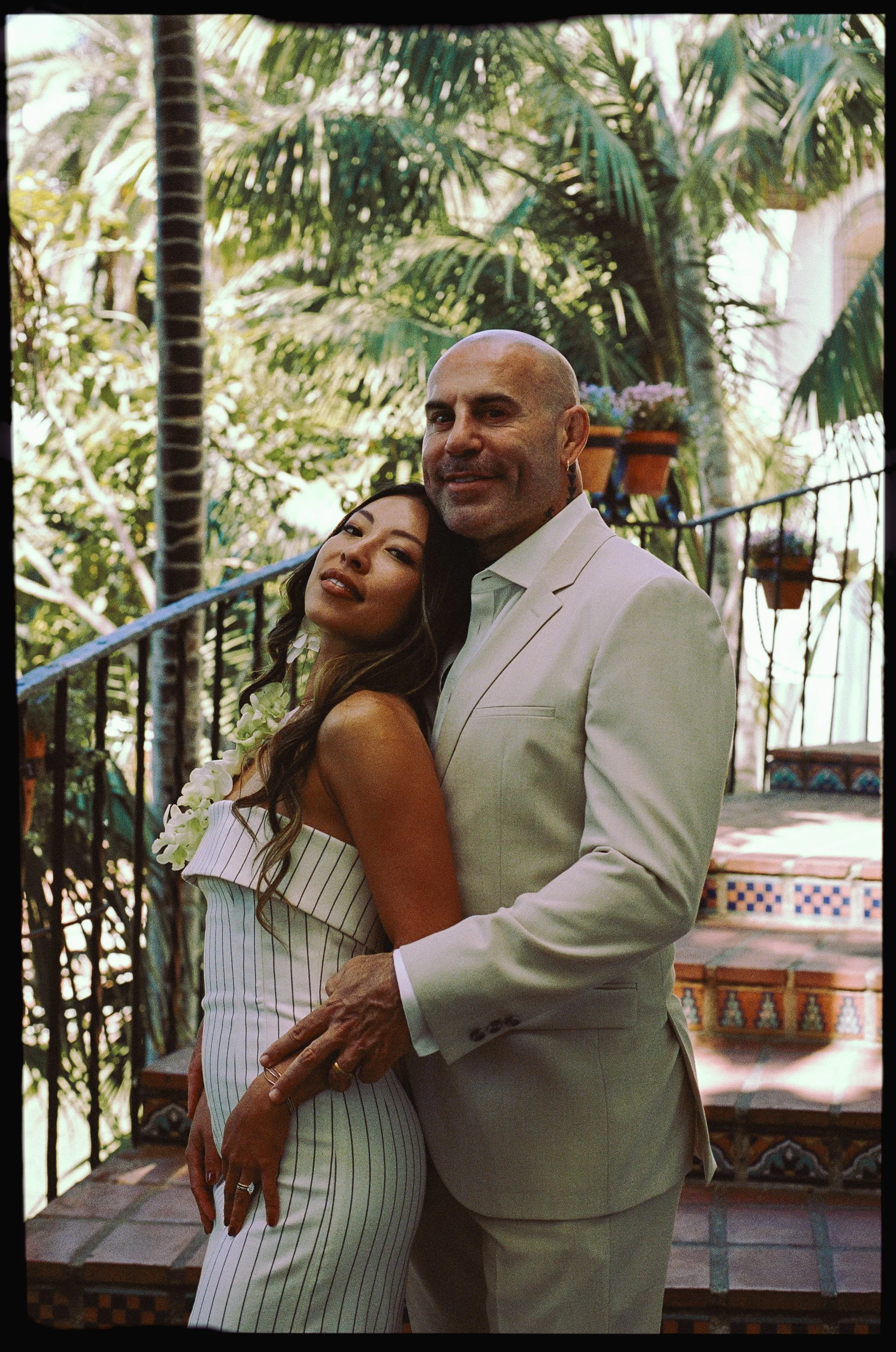A couple dressed in formal attire, standing closely together outdoors amid lush tropical plants and decorative tile steps.