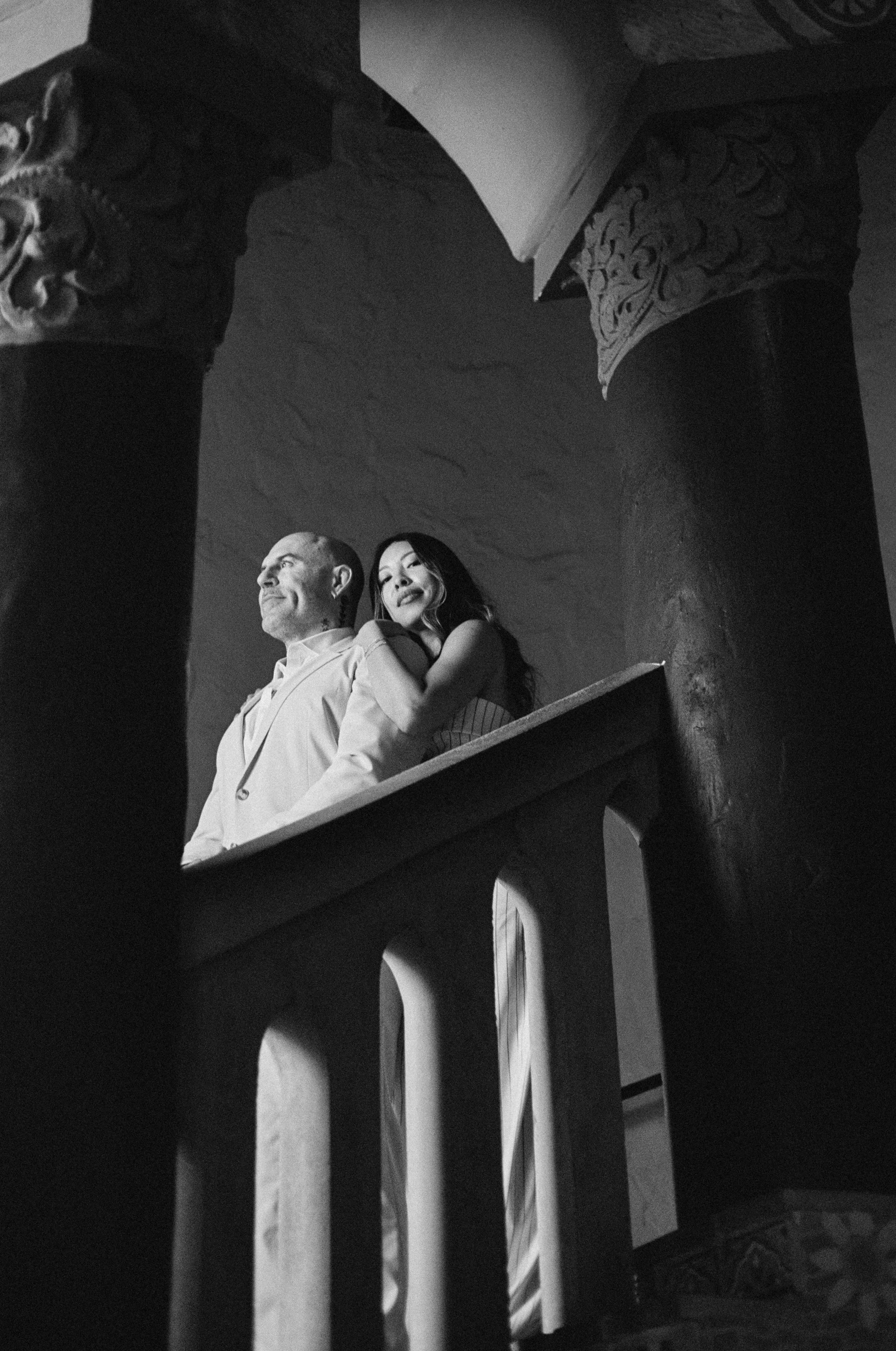 Black and white photograph of a man and a woman standing together on a balcony with ornate columns and railing, looking outward. The man is dressed in a suit, and the woman is wearing a sleeveless dress, both appearing thoughtful or contemplative.