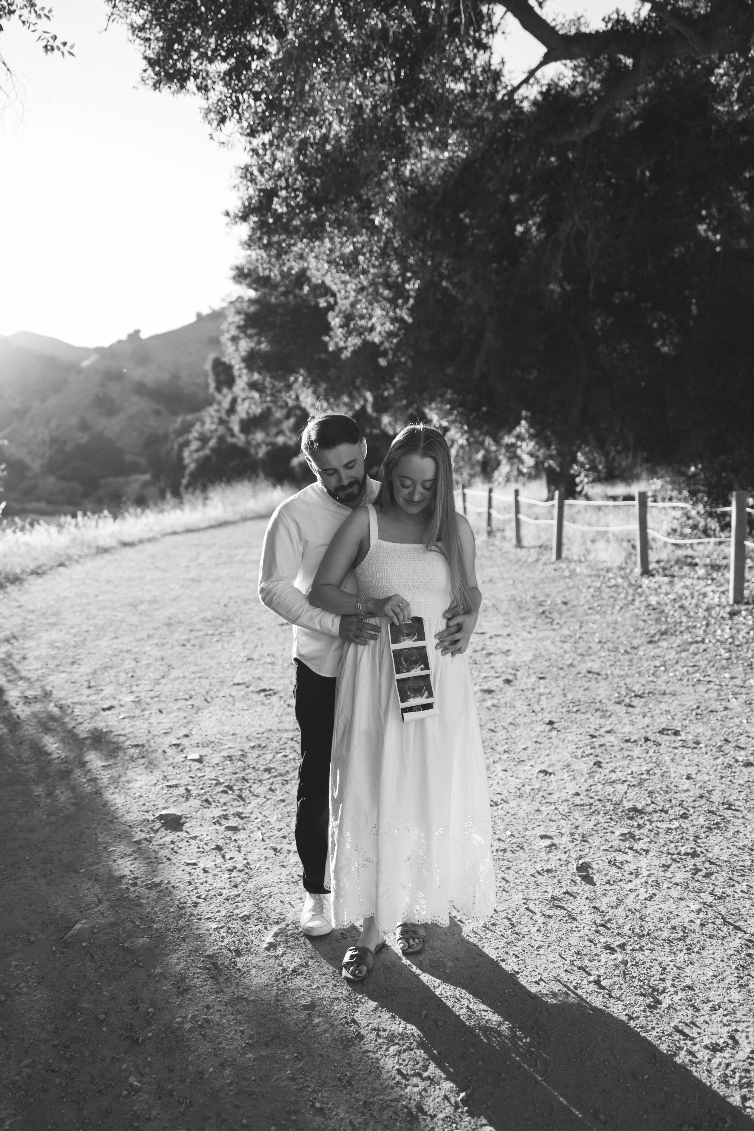 A couple walking outdoors on a dirt path, holding ultrasound images, with trees and hills in the background, captured in black and white.