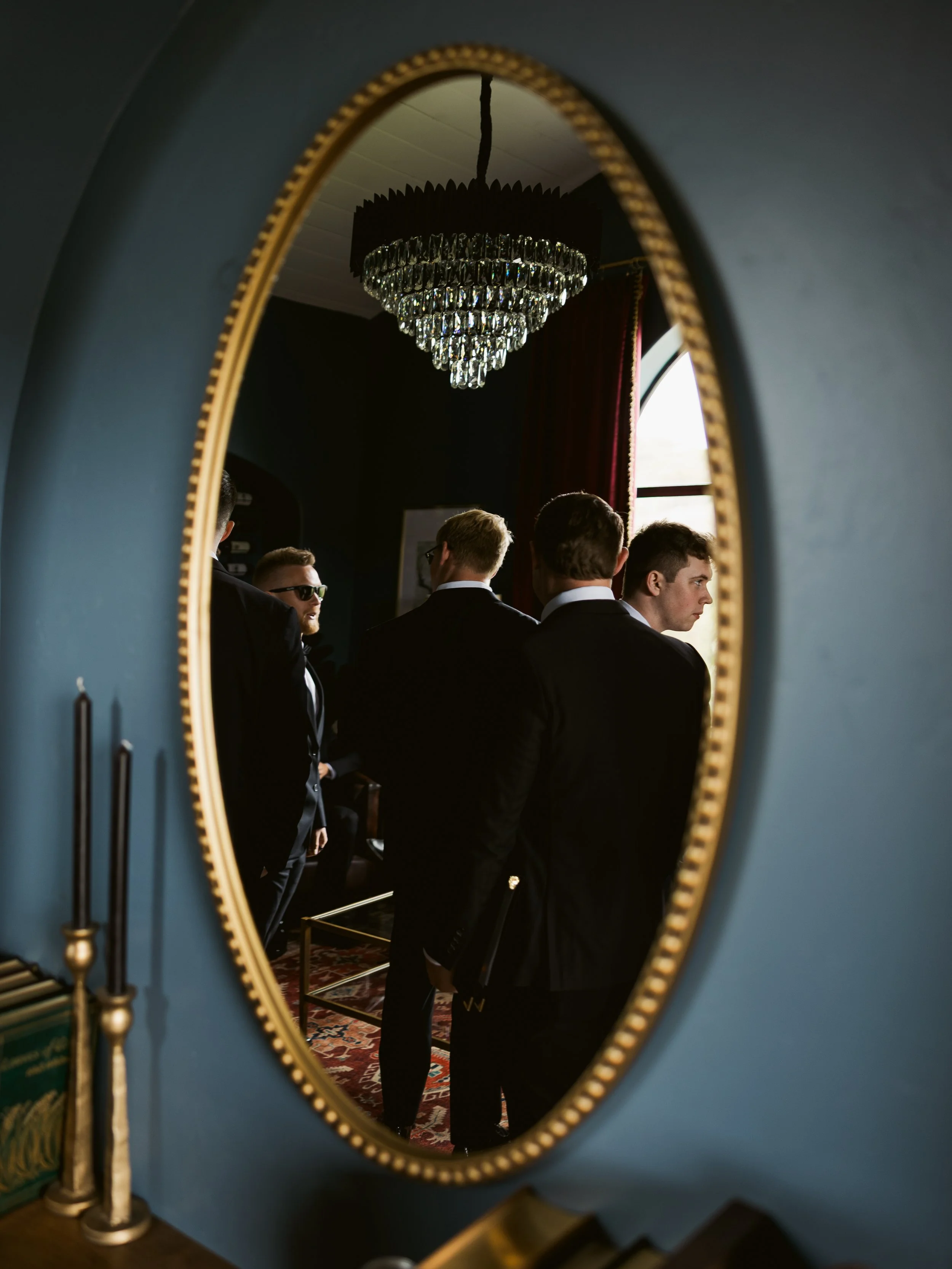 Reflection in an oval mirror showing a group of men dressed in black suits inside an elegant room with a chandelier and a window with red curtains.