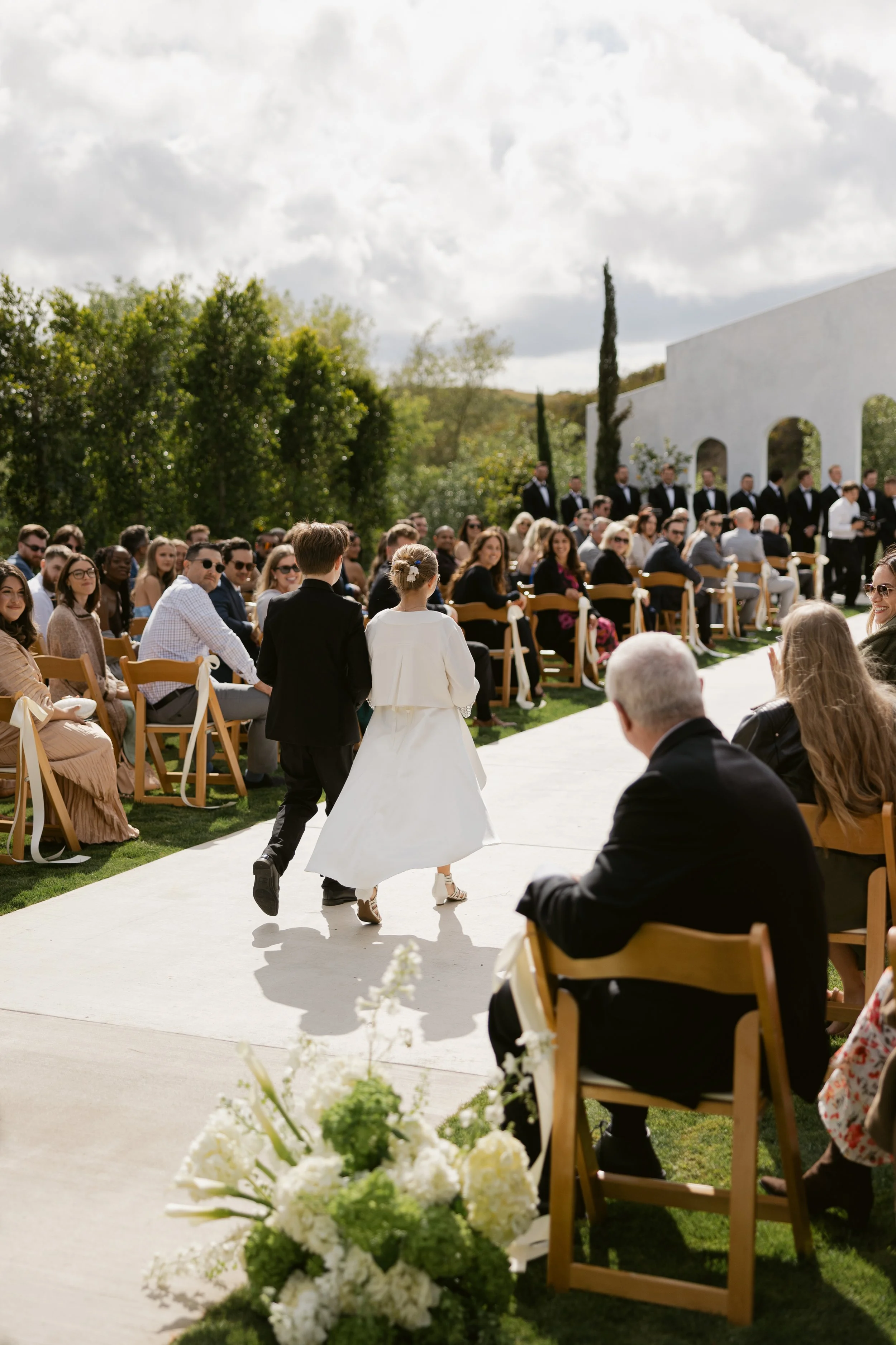 A bride and groom walking down the aisle at an outdoor wedding ceremony with guests seated on either side, greenery and trees in the background, under partly cloudy skies.