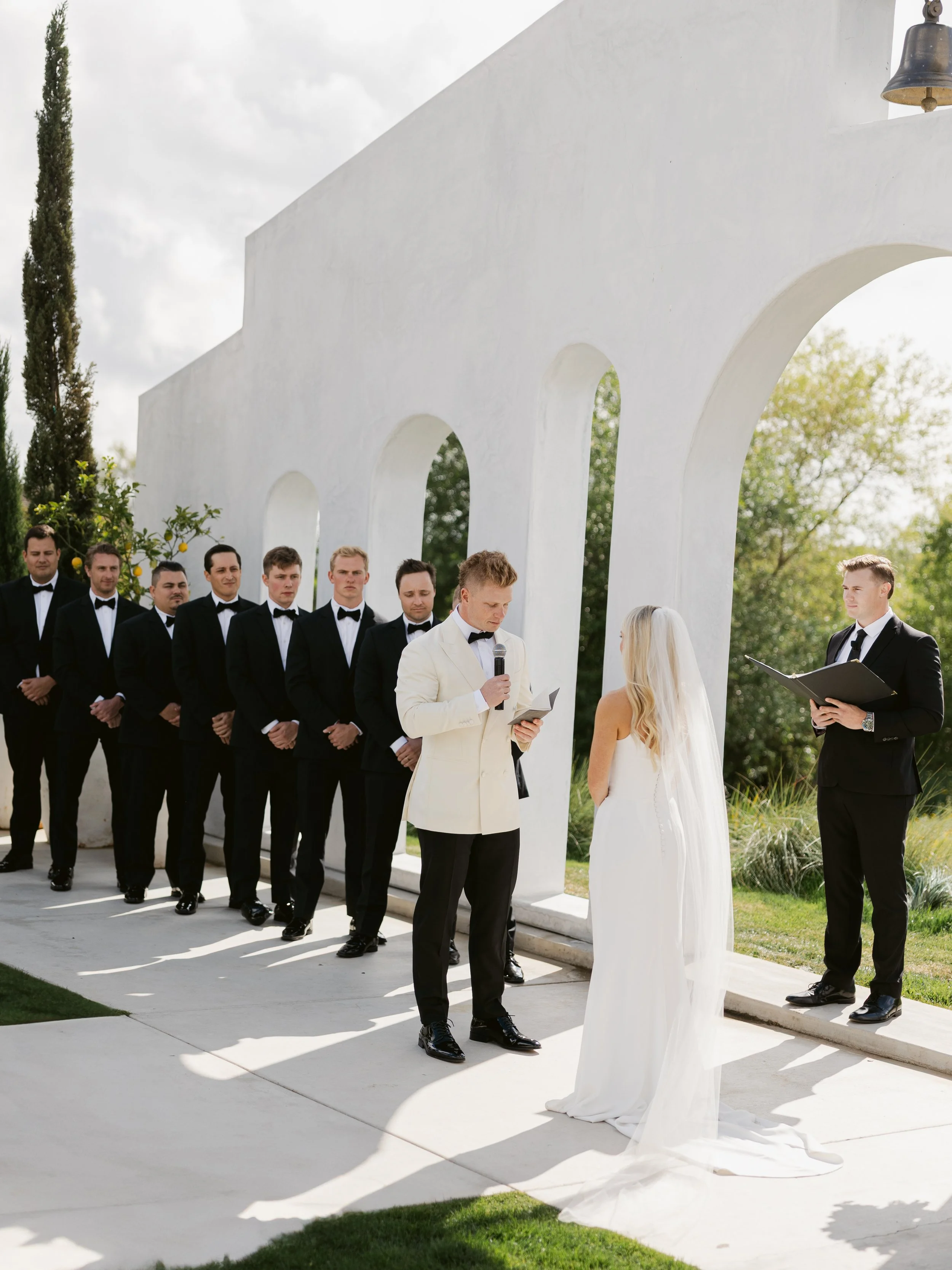 A wedding ceremony taking place outdoors with a bride, groom, and officiant, surrounded by groomsmen in black tuxedos, under white arches with greenery in the background.