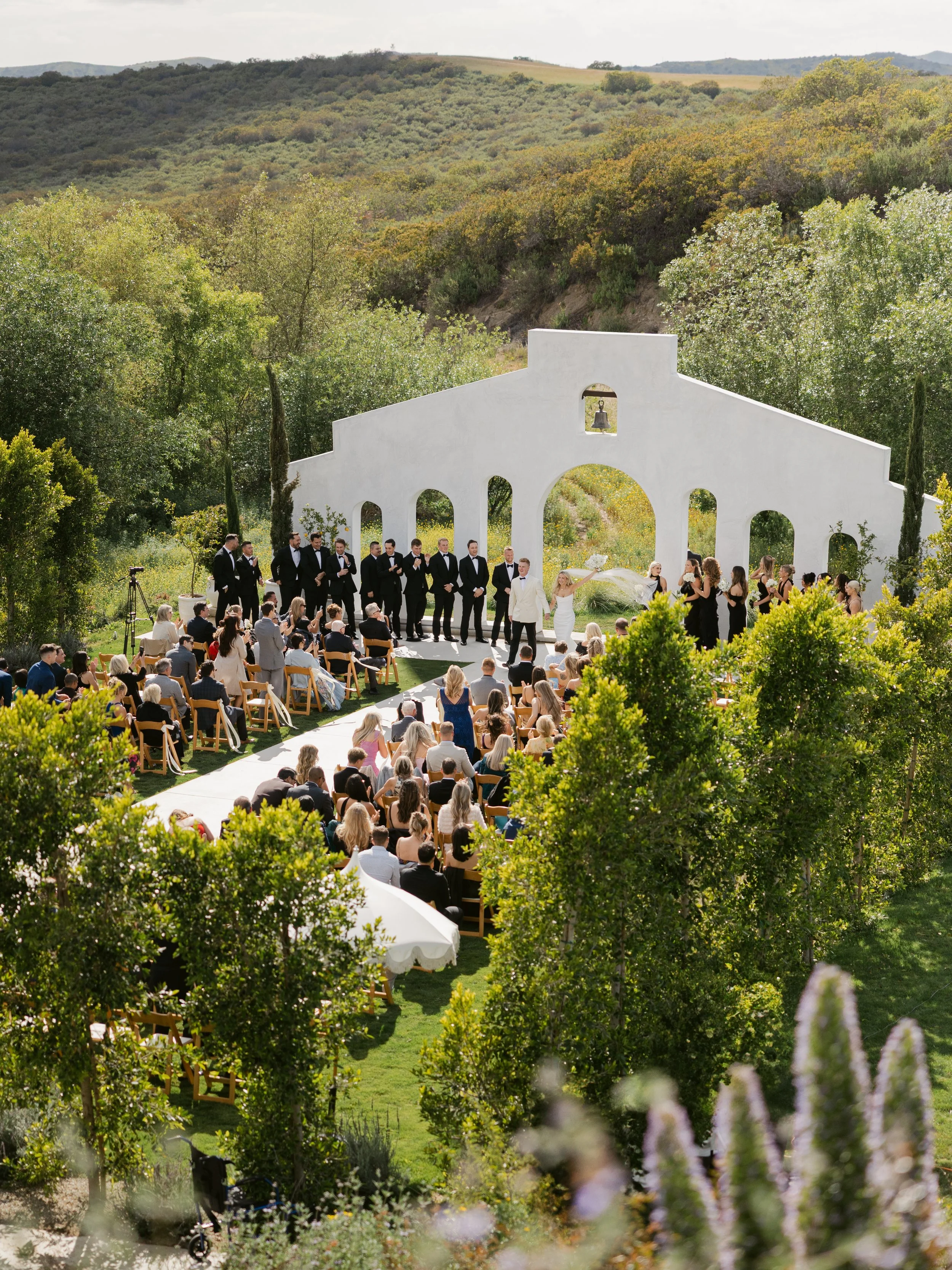 Outdoor wedding ceremony with guests seated on wooden chairs, surrounded by trees and green hills, featuring a white altar with a bell