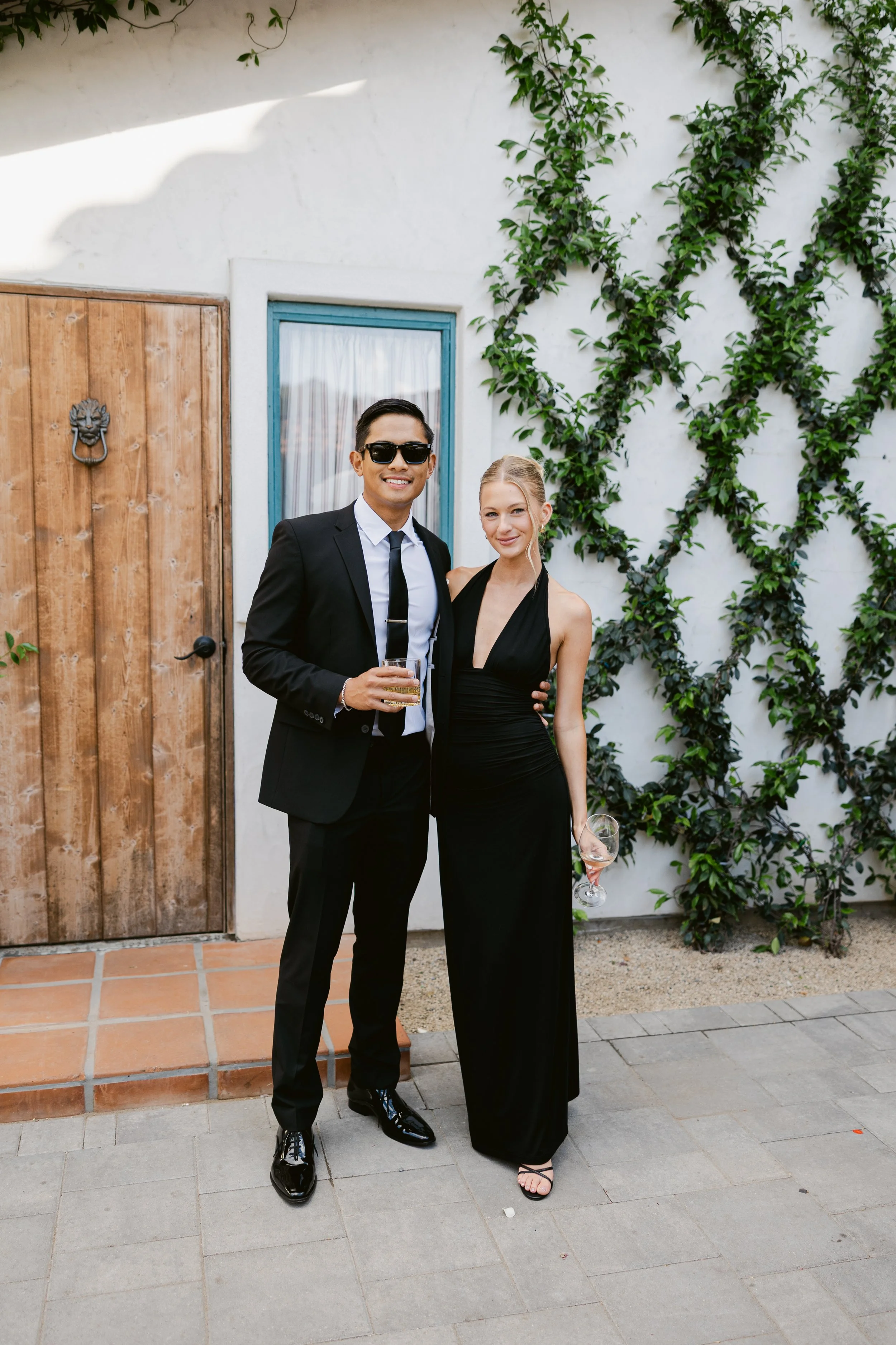 A man and woman dressed in formal black attire standing outside in front of a white wall with green vines. The man is wearing a black suit, white shirt, and black tie, with sunglasses, holding a drink. The woman is wearing a long black dress, holding