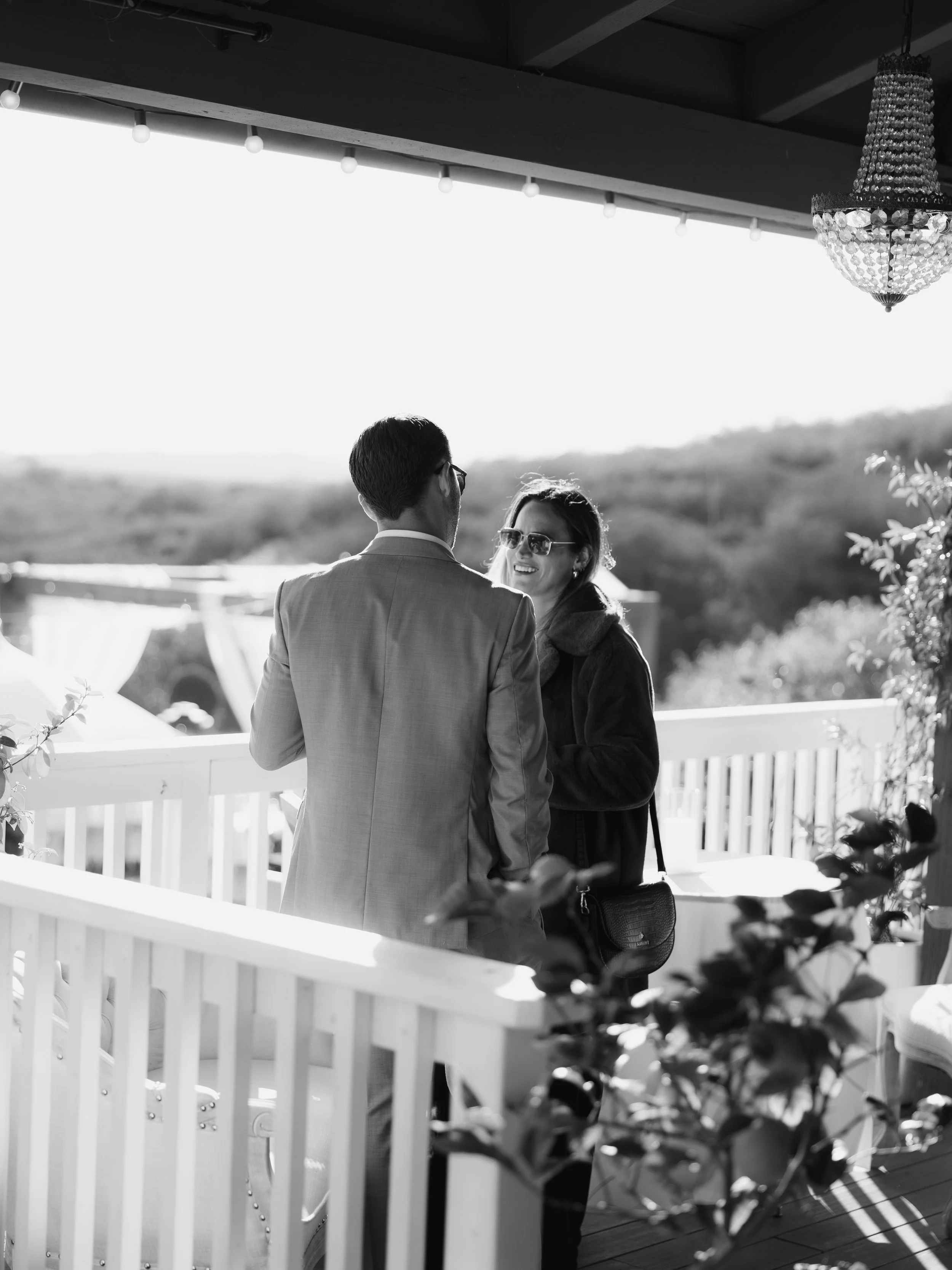 A man and a woman are talking on a balcony with scenic view in the background. The woman is smiling and wearing sunglasses, while the man has his back to the camera. There is a chandelier hanging from the ceiling and some plants in the foreground.