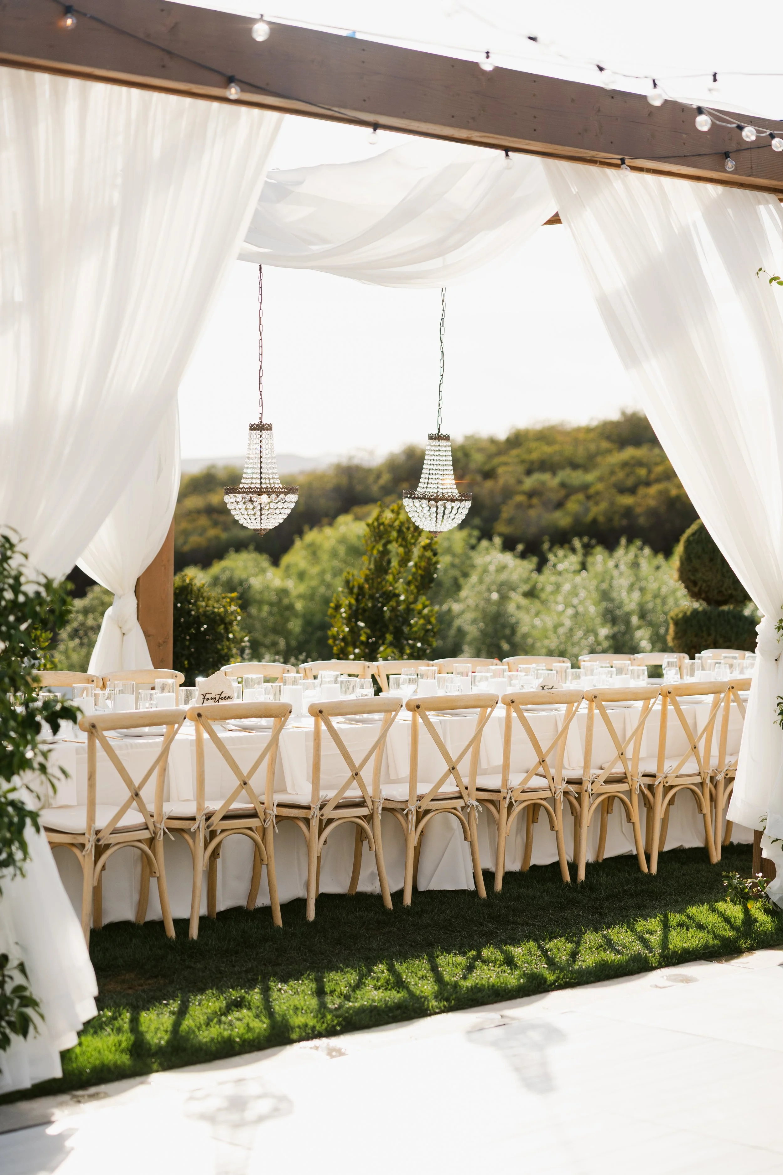 Elegant outdoor wedding reception setup with long table, beige cross-back chairs, white tablecloth, glassware, and two hanging chandeliers, under a white draped canopy with string lights, overlooking a green landscape.