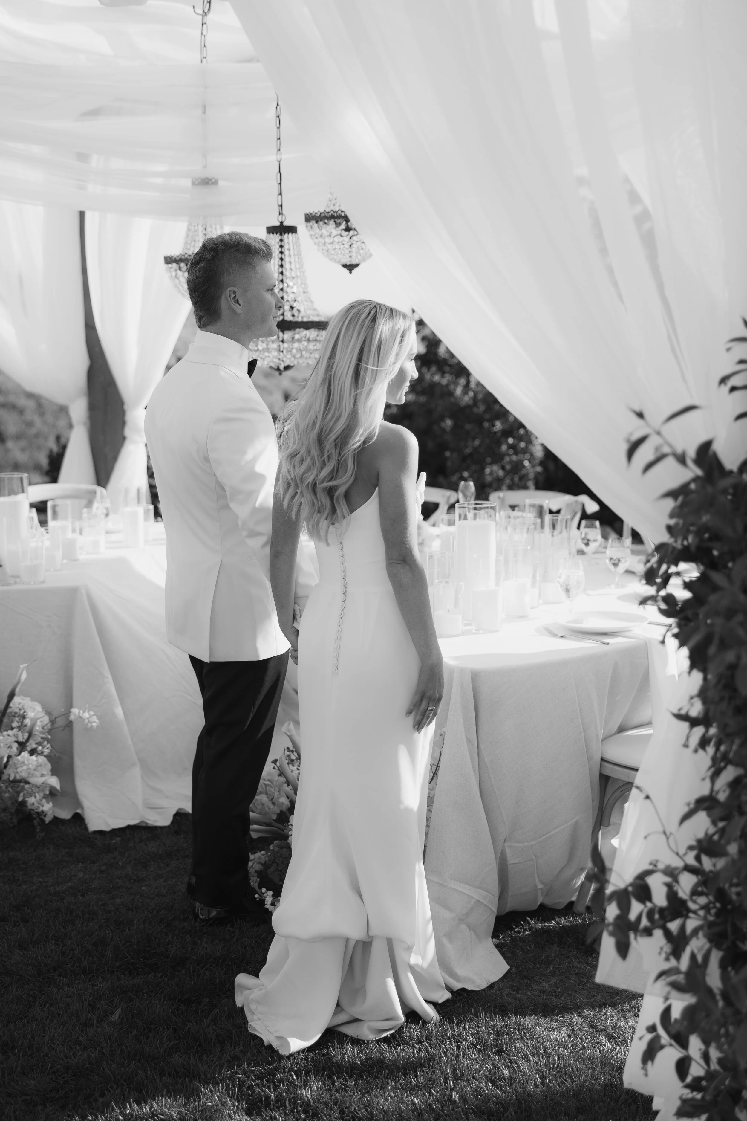 Black and white photo of a bride and groom standing hand-in-hand during their wedding ceremony outdoors, with elegant drapery and chandeliers overhead, and a decorated banquet table nearby.