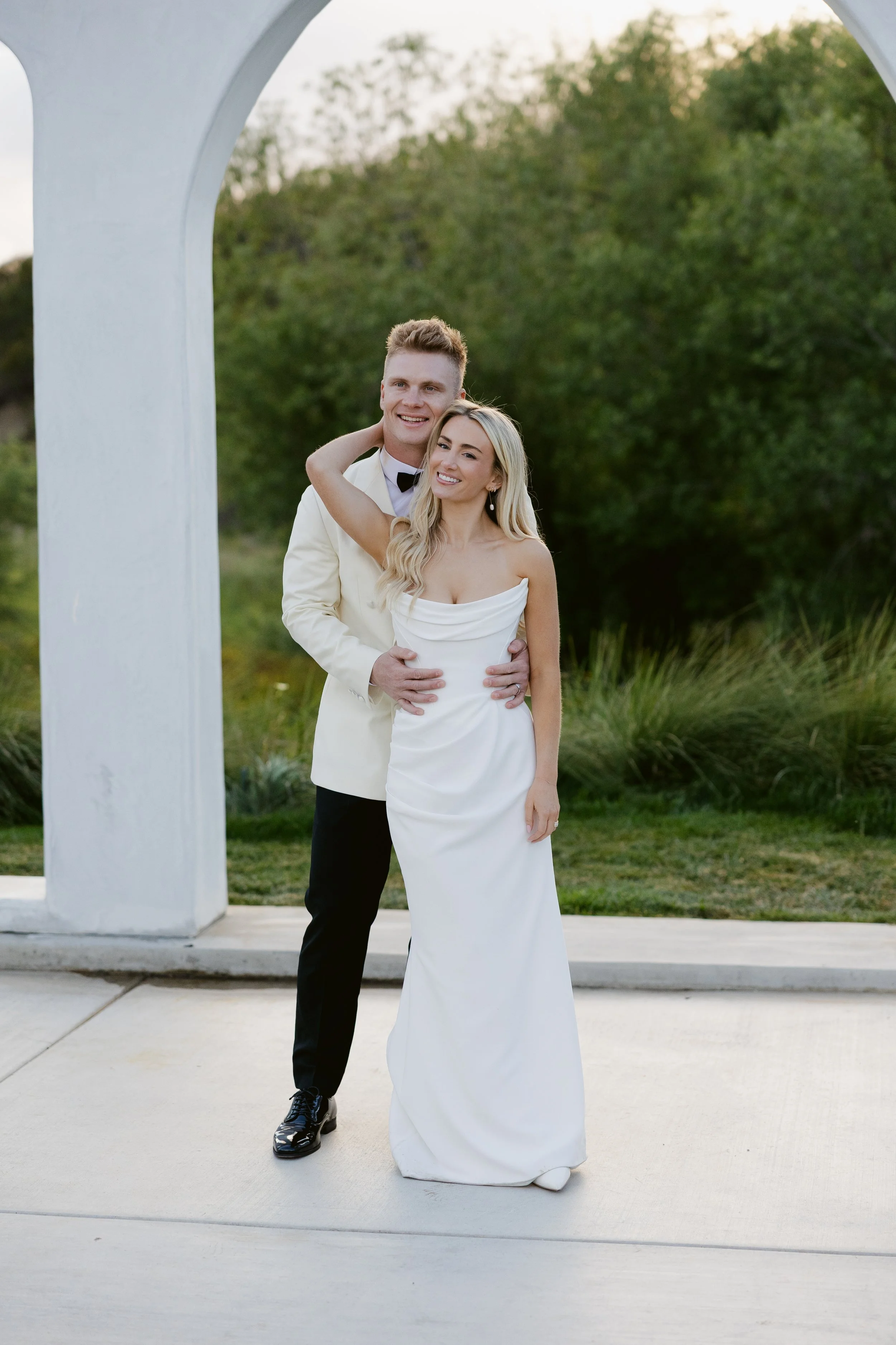 A newlywed couple dressed in wedding attire, smiling and standing outdoors under a white arch, with greenery in the background.