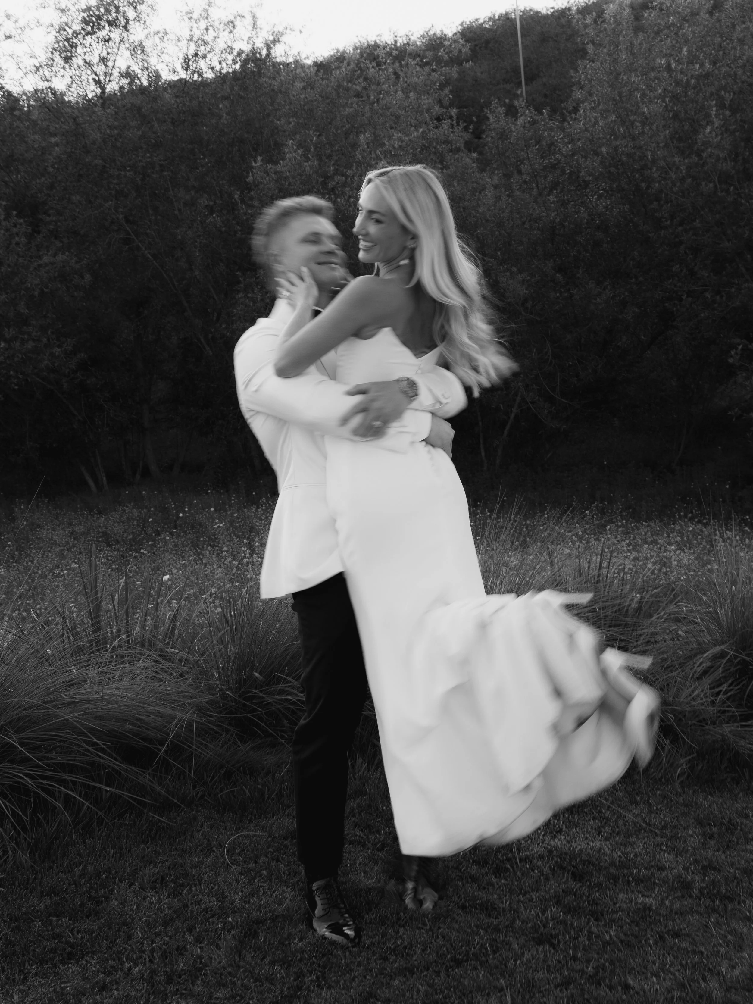 A black and white photograph of a man lifting a woman in a white dress outdoors, with trees and grass in the background.