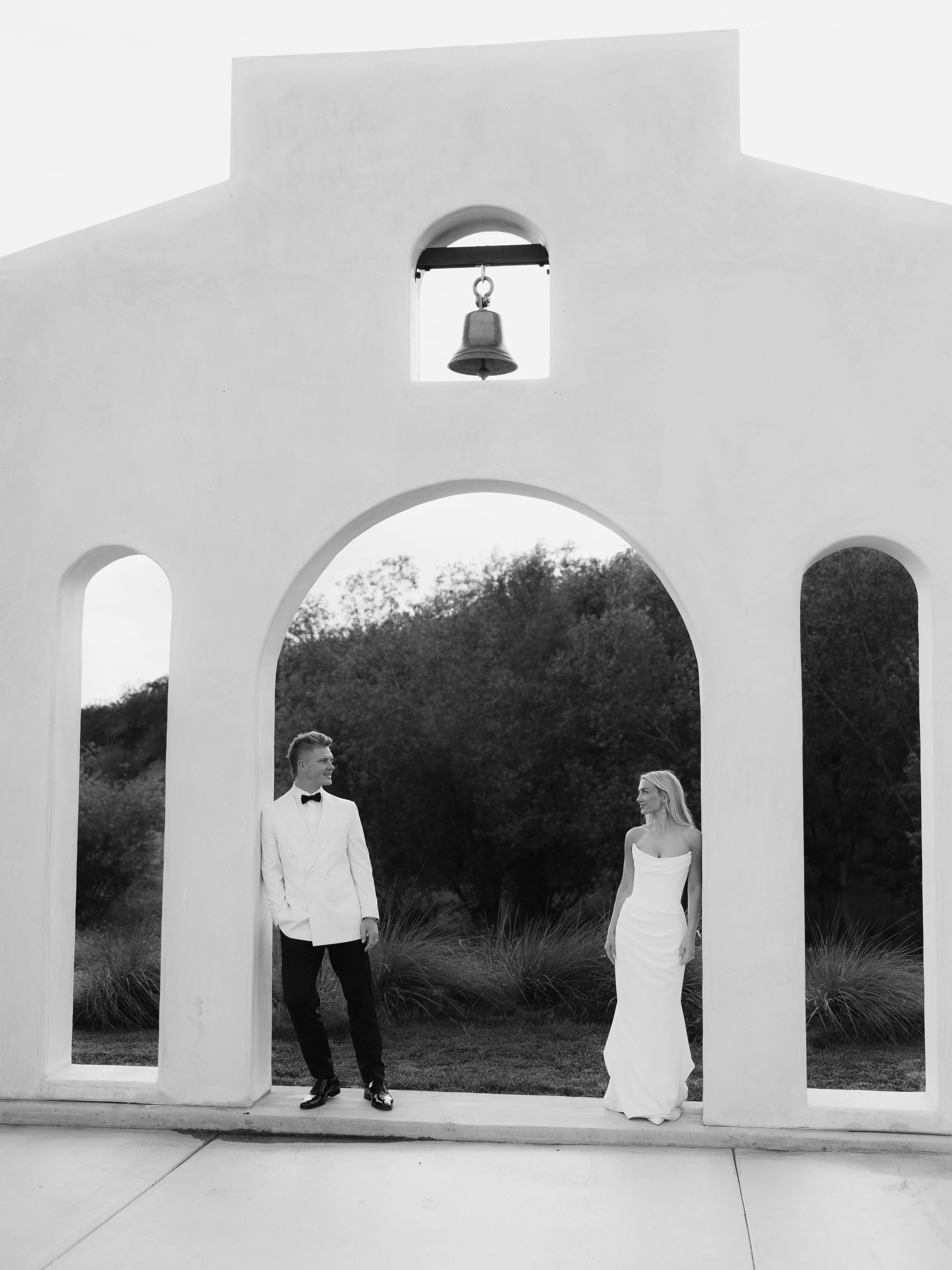 A black and white photo of a bride and groom standing in front of a white church with a bell tower, trees in the background, and a curved archway.