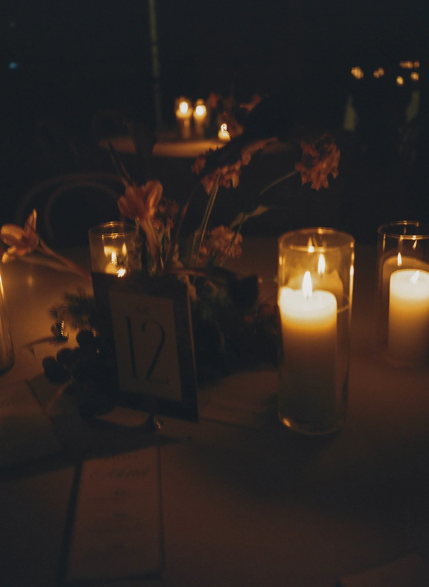 Dimly lit scene with lit candles on a table, flowers, and blurred background lights.