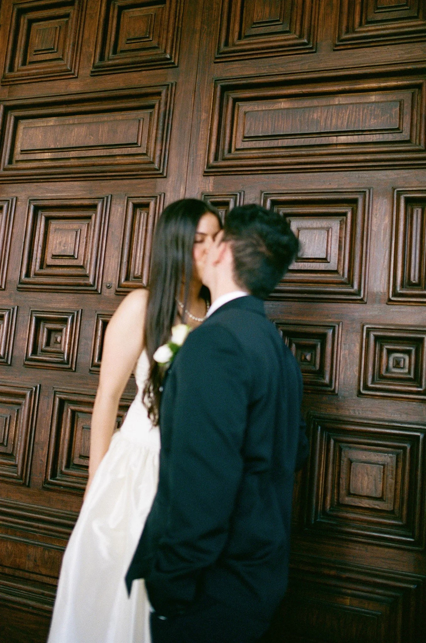 A couple dressed in wedding attire, sharing a kiss in front of a large, ornate wood-paneled wall.