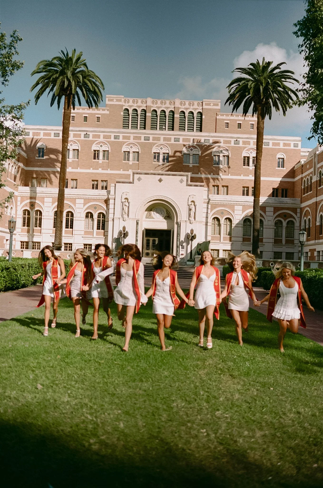 Group of young women in white dresses and orange sashes walking on green grass in front of a large historic building with palm trees.