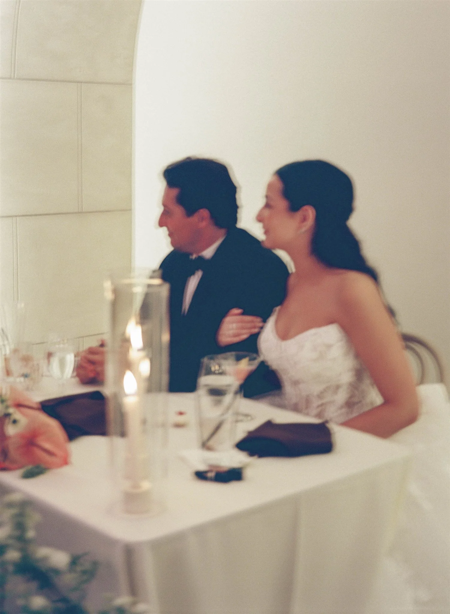 A man and woman sit at a table during a formal event, likely a wedding, with candles and flowers on the table.