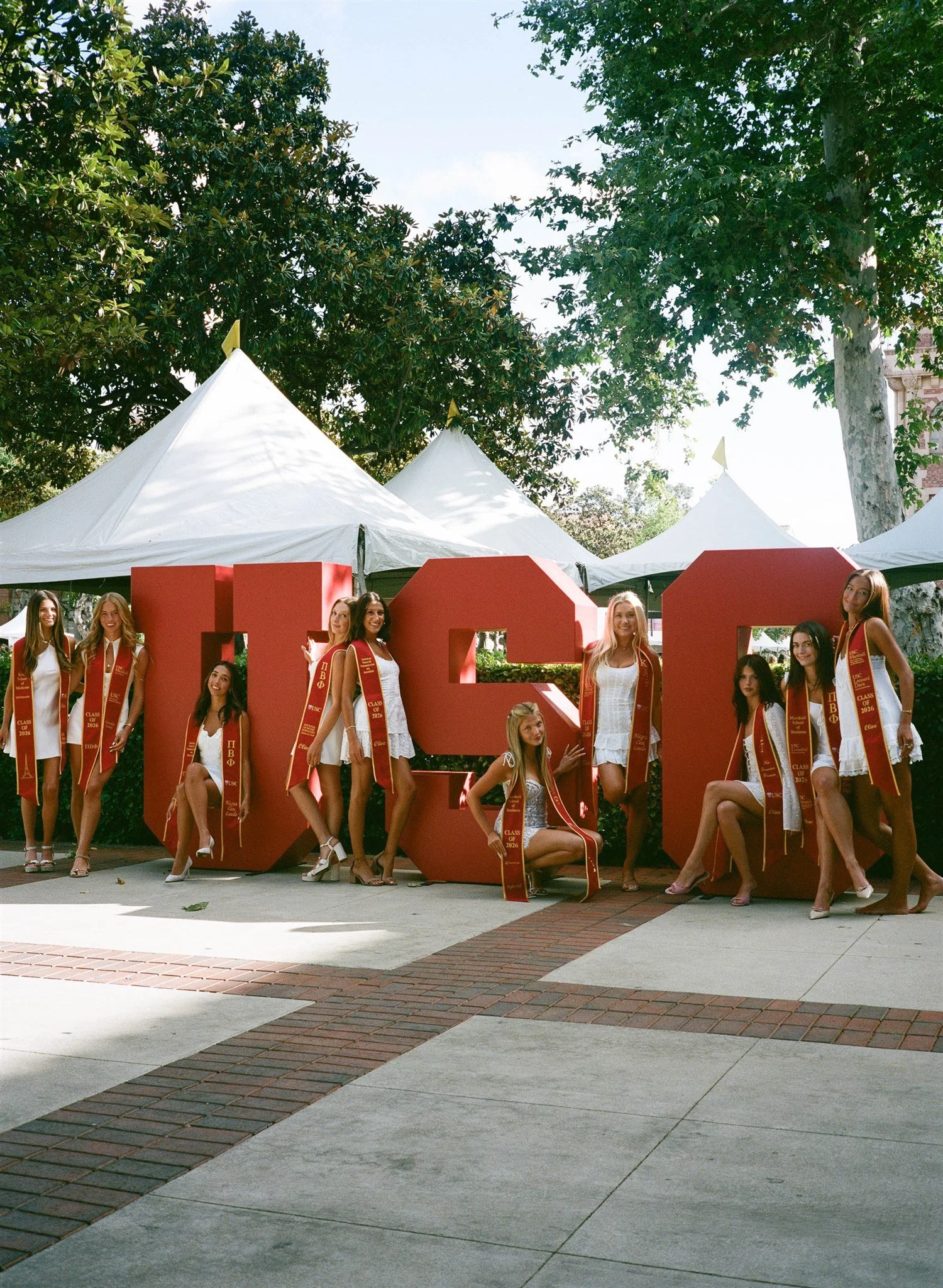 A group of young women wearing graduation caps and robes with red and gold sashes, posing in front of large red letters spelling 'USC', outdoors with trees and tents in the background.