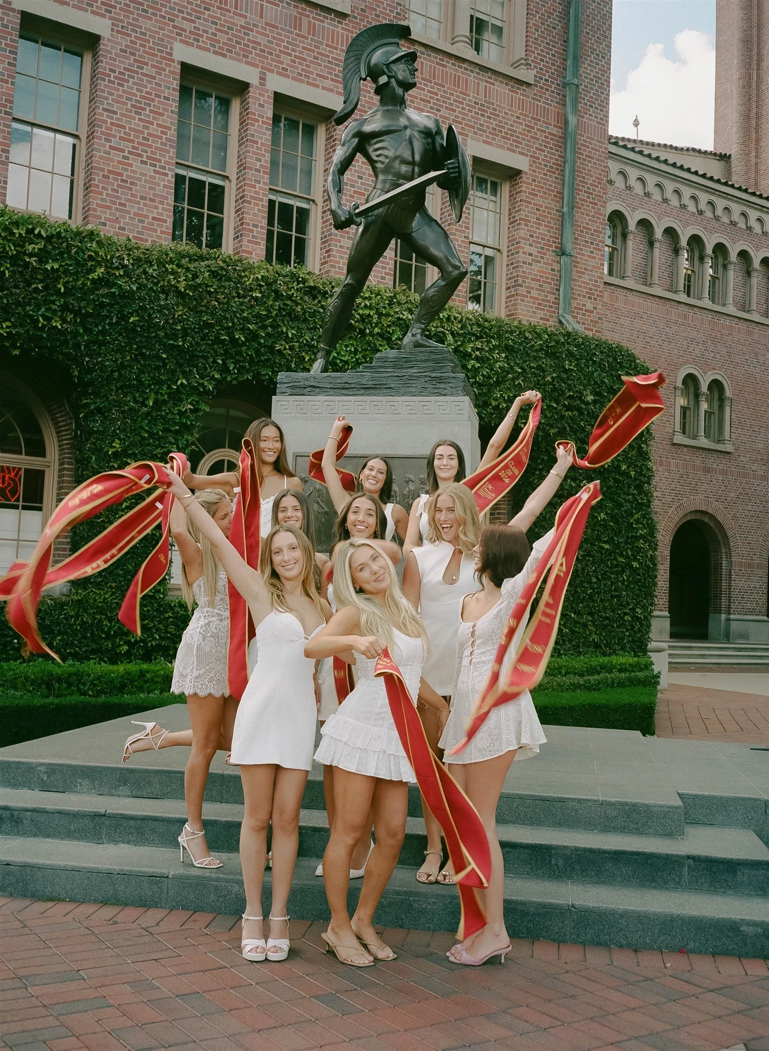A group of young women in white dresses celebrating in front of a statue of a Roman soldier, holding red and gold ribbons, on steps outside a brick building.