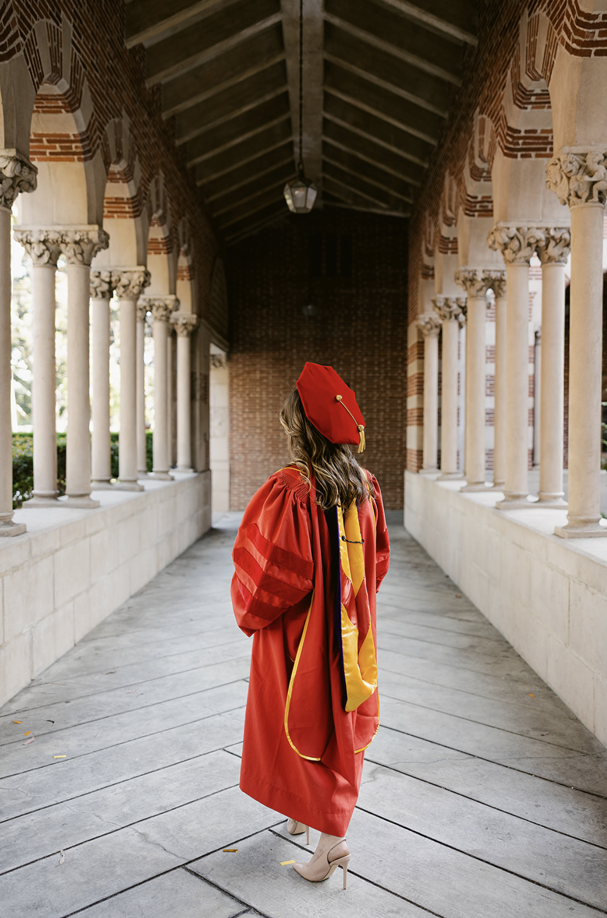 Woman in graduation gown and cap standing in a colonnade archway with brick and stone walls.