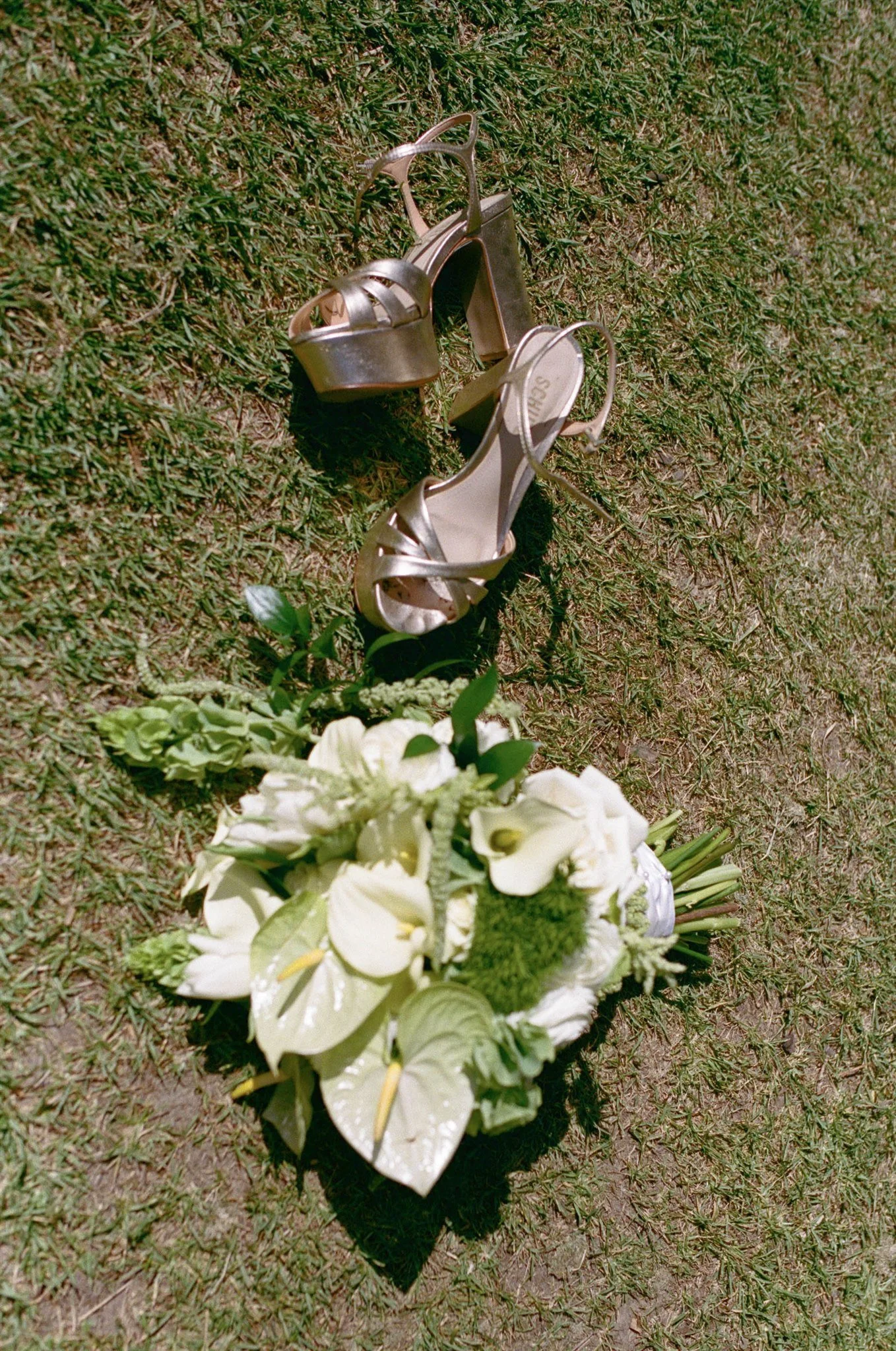 A pair of silver high-heeled sandals on a grassy surface, accompanied by a white floral arrangement.