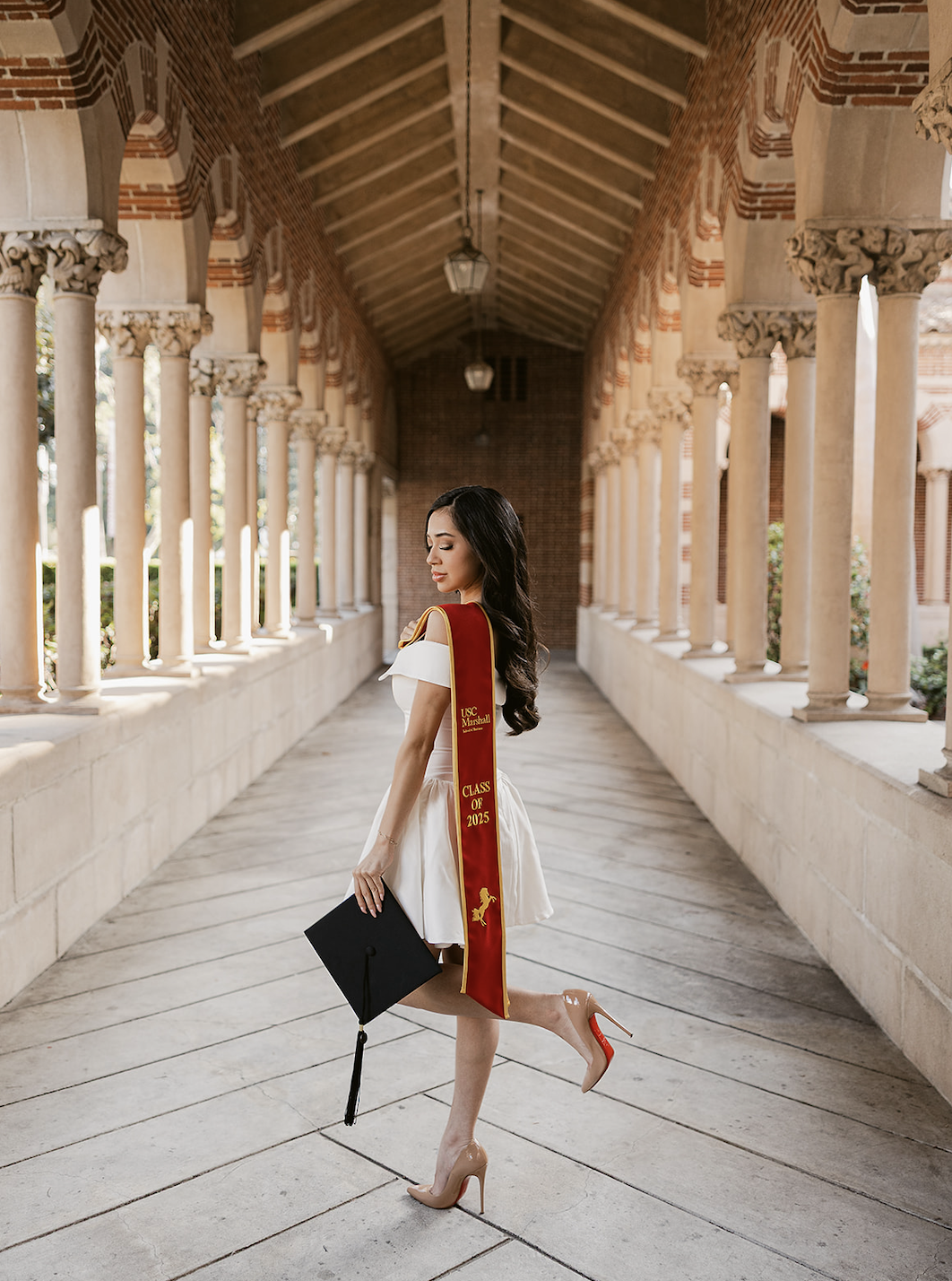 A young woman in a white dress and nude high heels is standing in a colonnade, holding a graduation cap and wearing a maroon and gold graduation stole with the words "USC Marshall, Class of 2025" and a yellow horse emblem.
