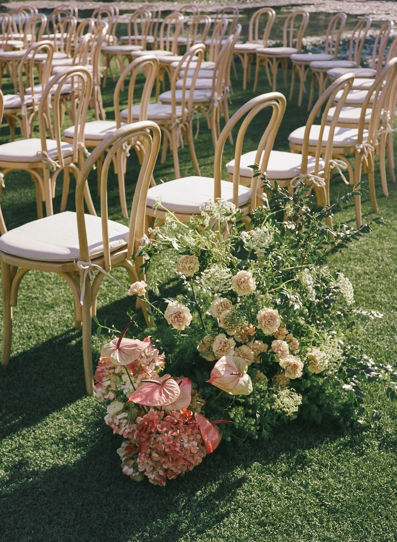Multiple wooden chairs with white cushions arranged outdoors near a body of water, with a floral arrangement of pink, white, and green flowers in the foreground.