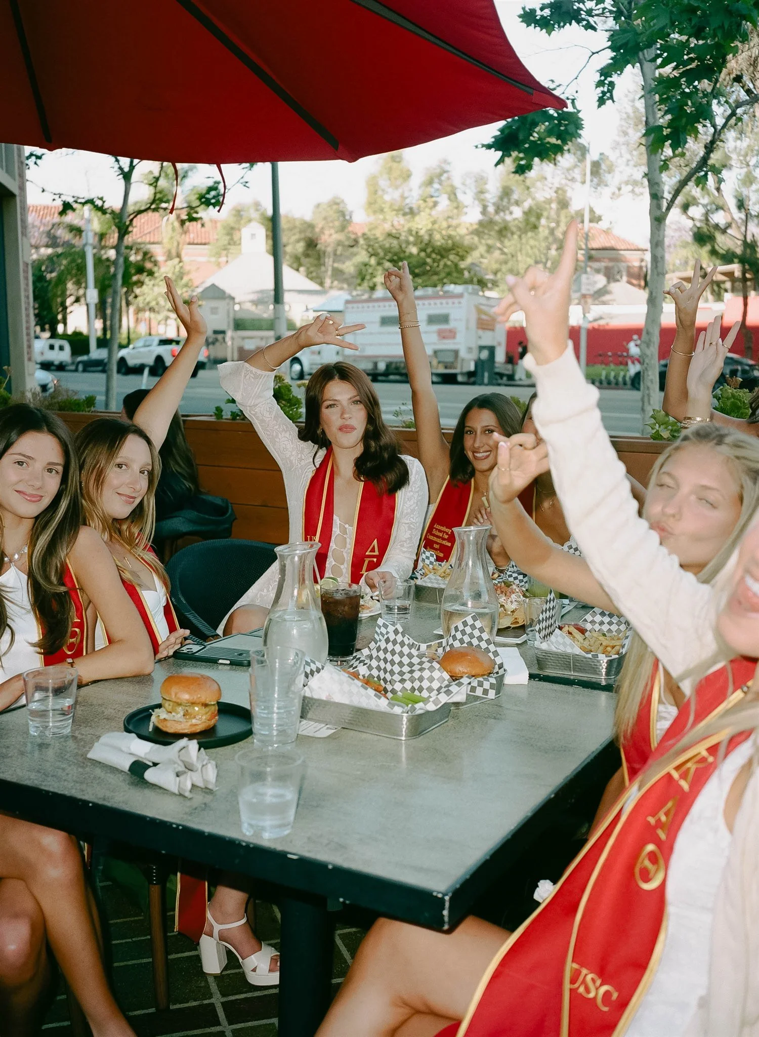 A group of young women wearing sorority sashes are sitting around a table outside at a restaurant, raising their hands in celebration. The table has sandwiches, drinks, and checkered paper food containers.