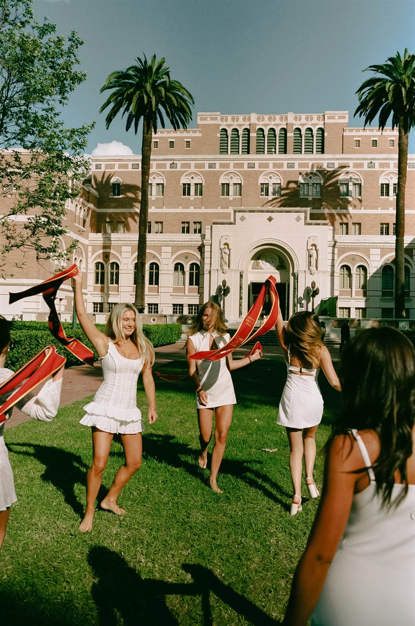 Young women in white dresses celebrating outdoors in front of a historic building with palm trees, holding red ribbons.