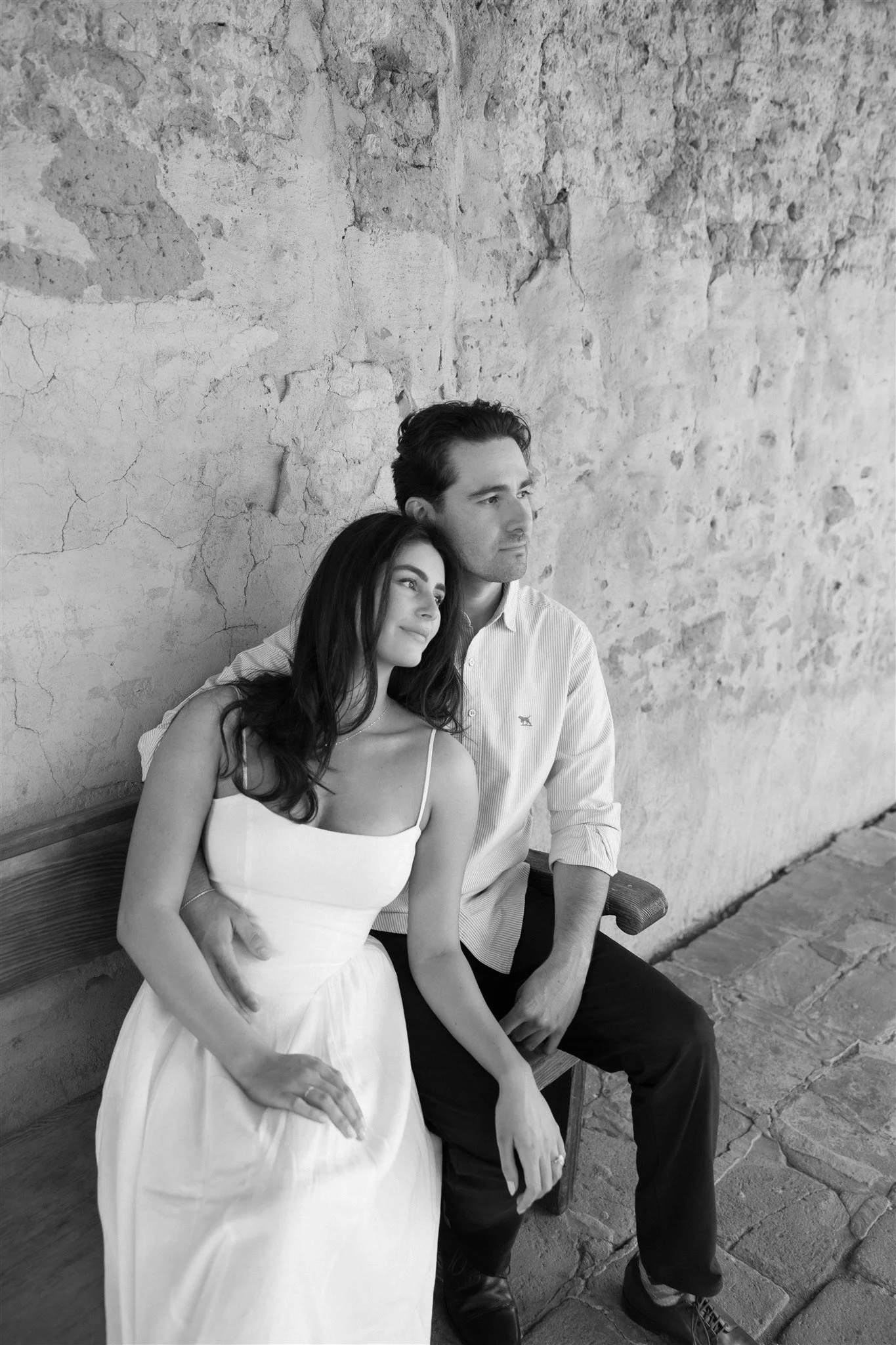 A black and white photo of a woman and a man sitting on a bench against a textured, cracked wall. The woman is wearing a white dress and leaning her head on the man's shoulder, who is wearing a light-colored shirt and dark pants.