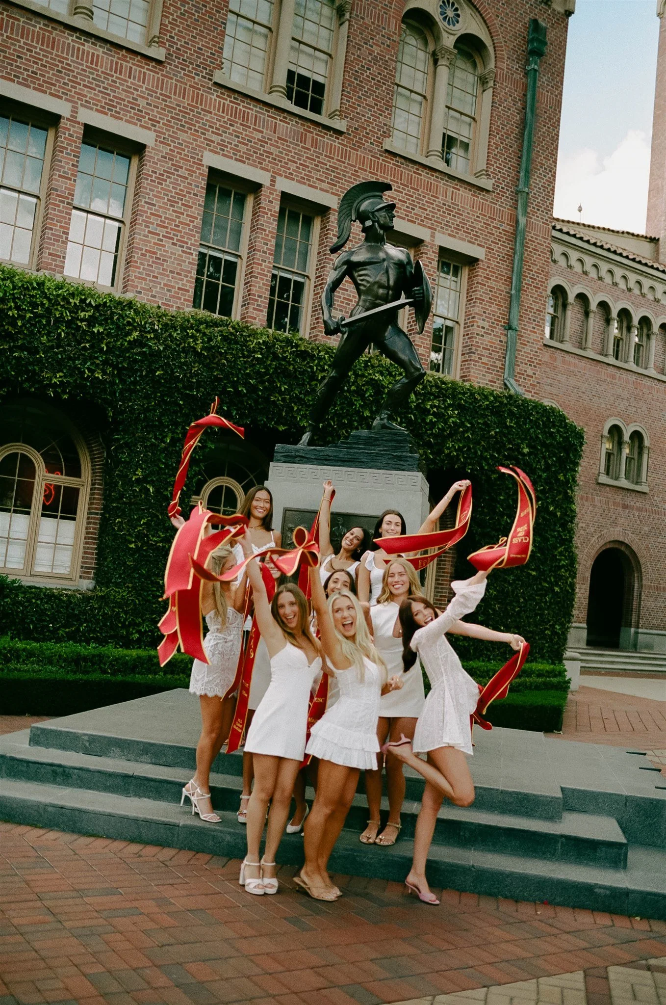 Group of women in white dresses celebrating on steps in front of a statue of a warrior in a helmet, with a brick building in the background.