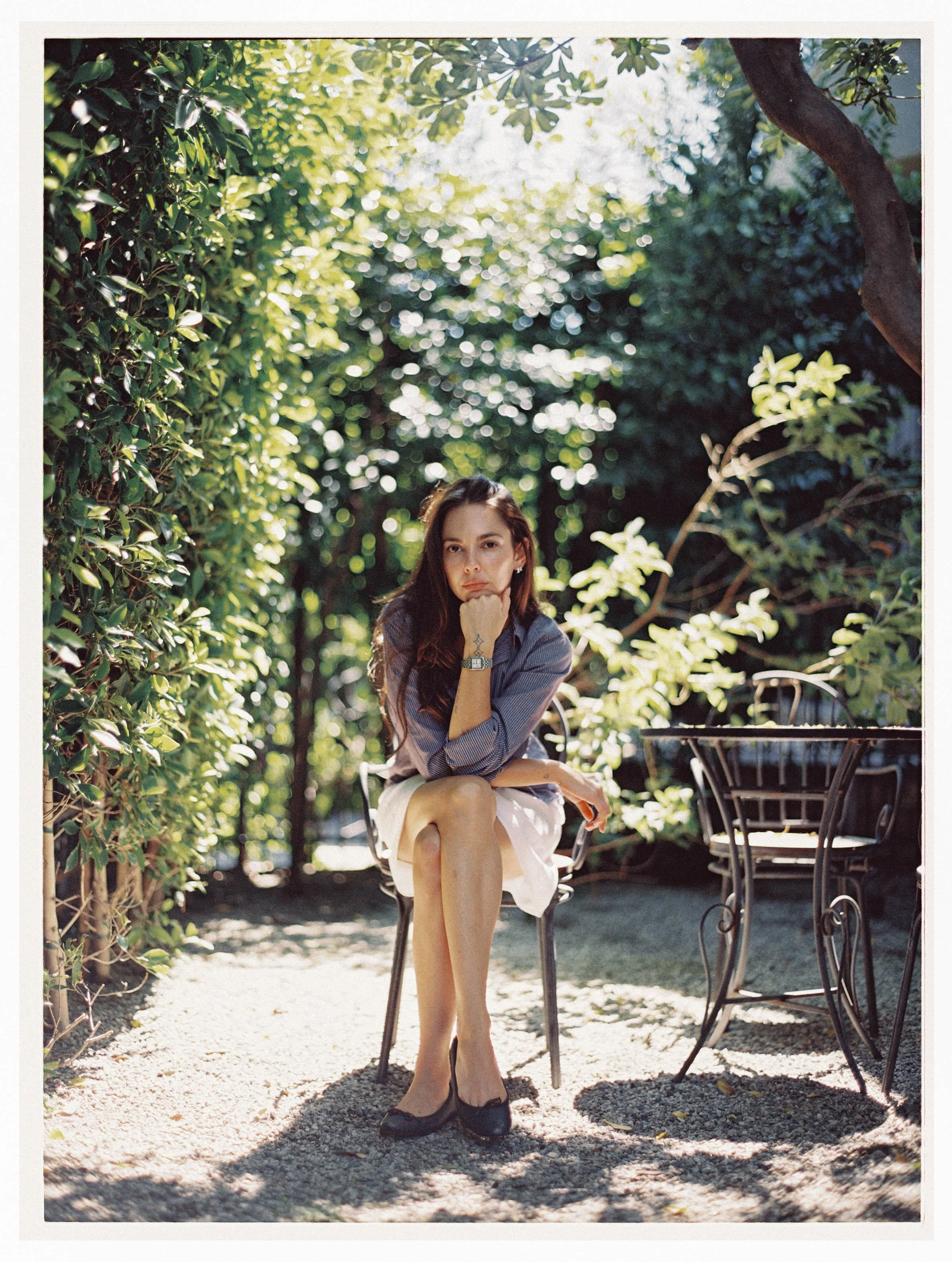 A woman with long brown hair and a serious expression, sitting outdoors on a black metal chair, resting her chin on her hand, surrounded by lush green foliage and dappled sunlight.