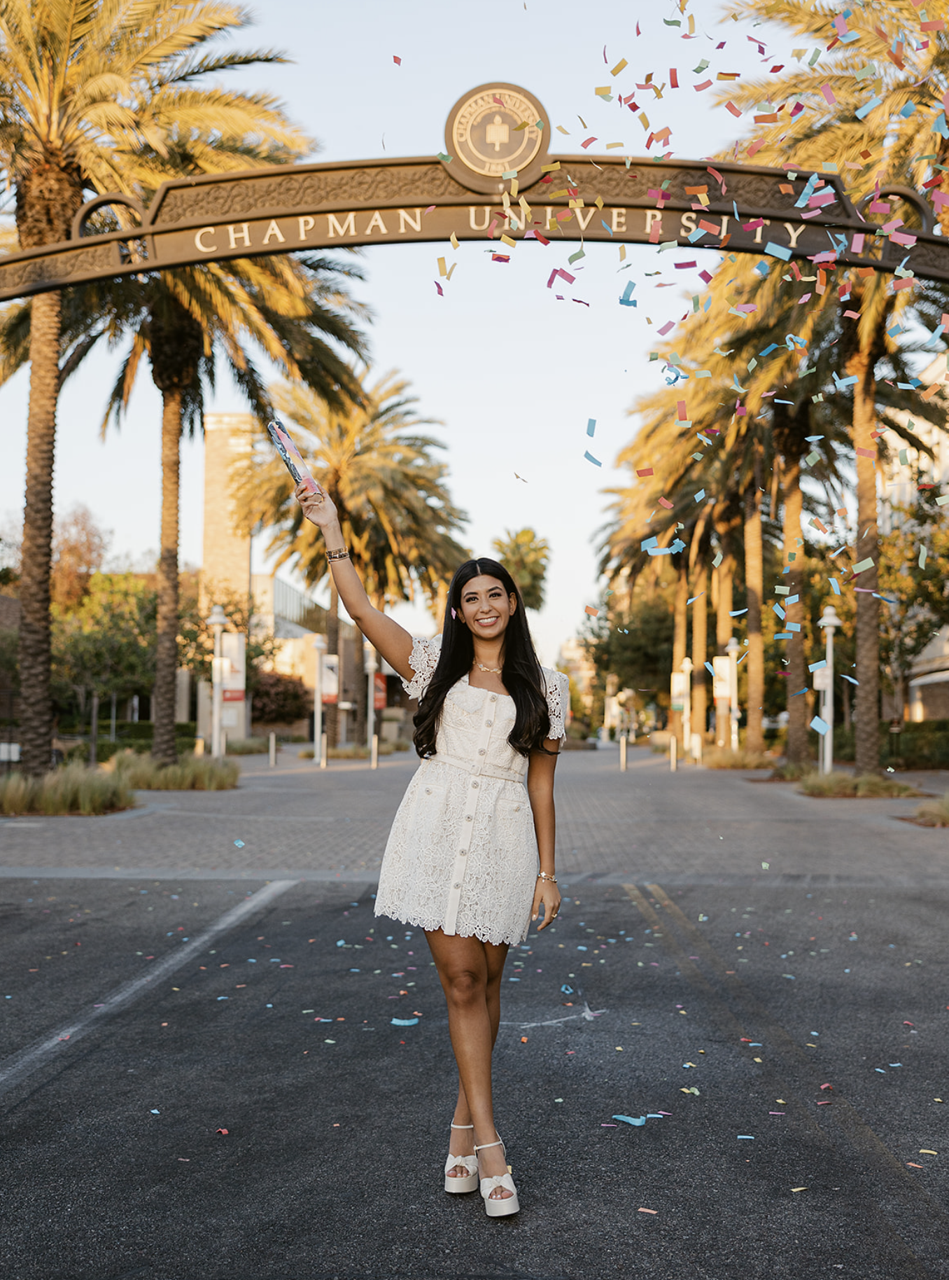 A woman celebrating graduation at Chapman University, standing on a parking lot with palm trees in the background, throwing colorful confetti in the air, wearing a white lace dress and high-heeled shoes.