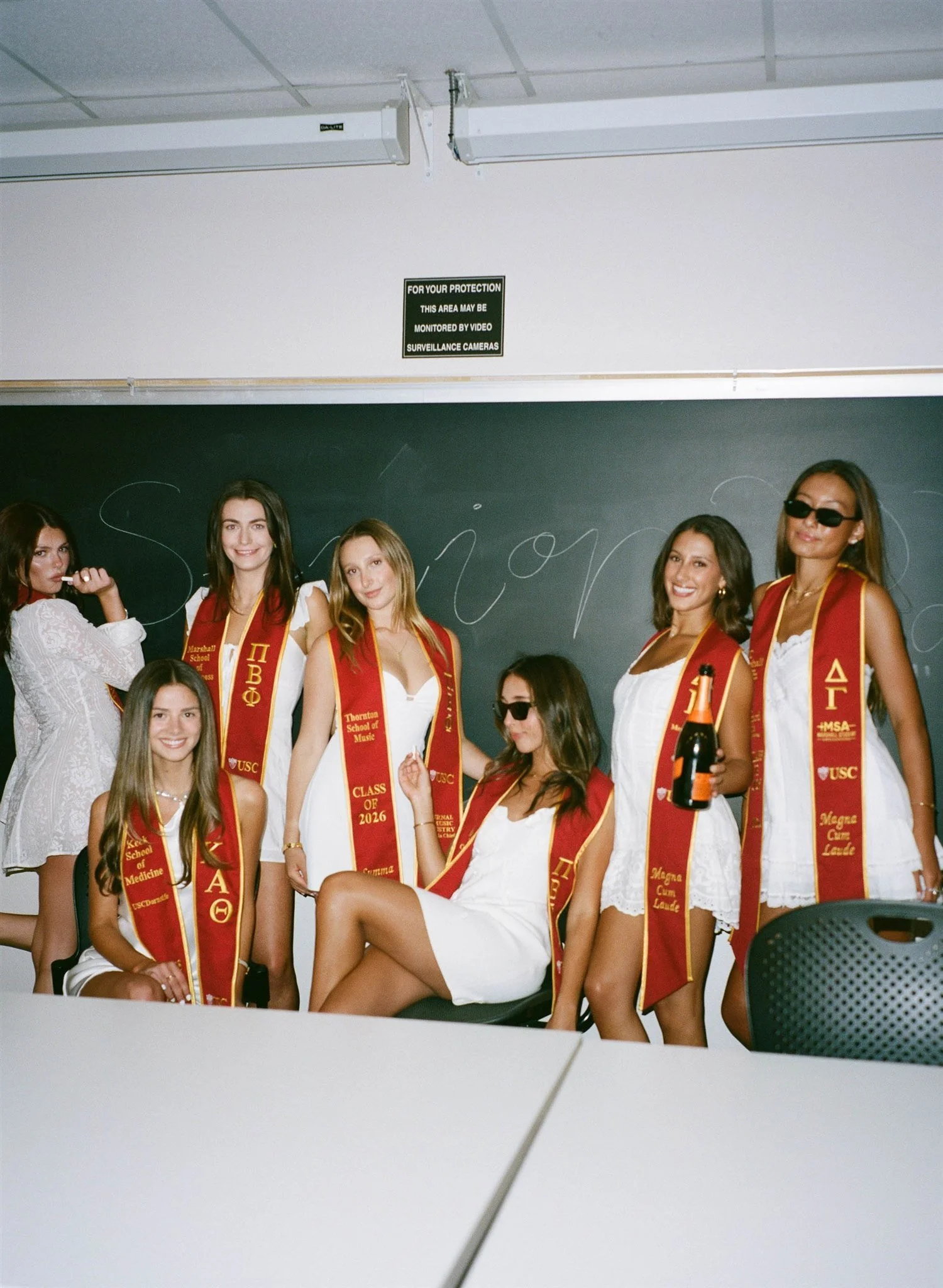 Group of young women in white dresses wearing red and gold sashes, some smiling and posing, in a classroom with a blackboard and a sign that reads 'For your protection, this area may be monitored by video surveillance cameras'
