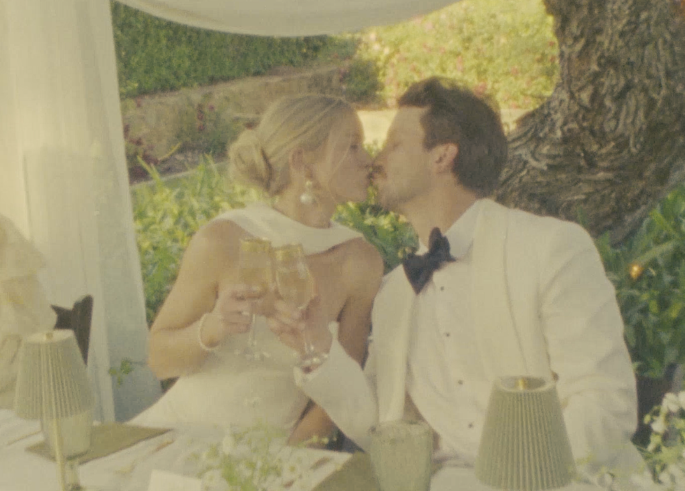 A couple dressed in wedding attire, sharing a kiss outdoors, with glasses of champagne in hand, sitting at a decorated table with a tree and greenery in the background.