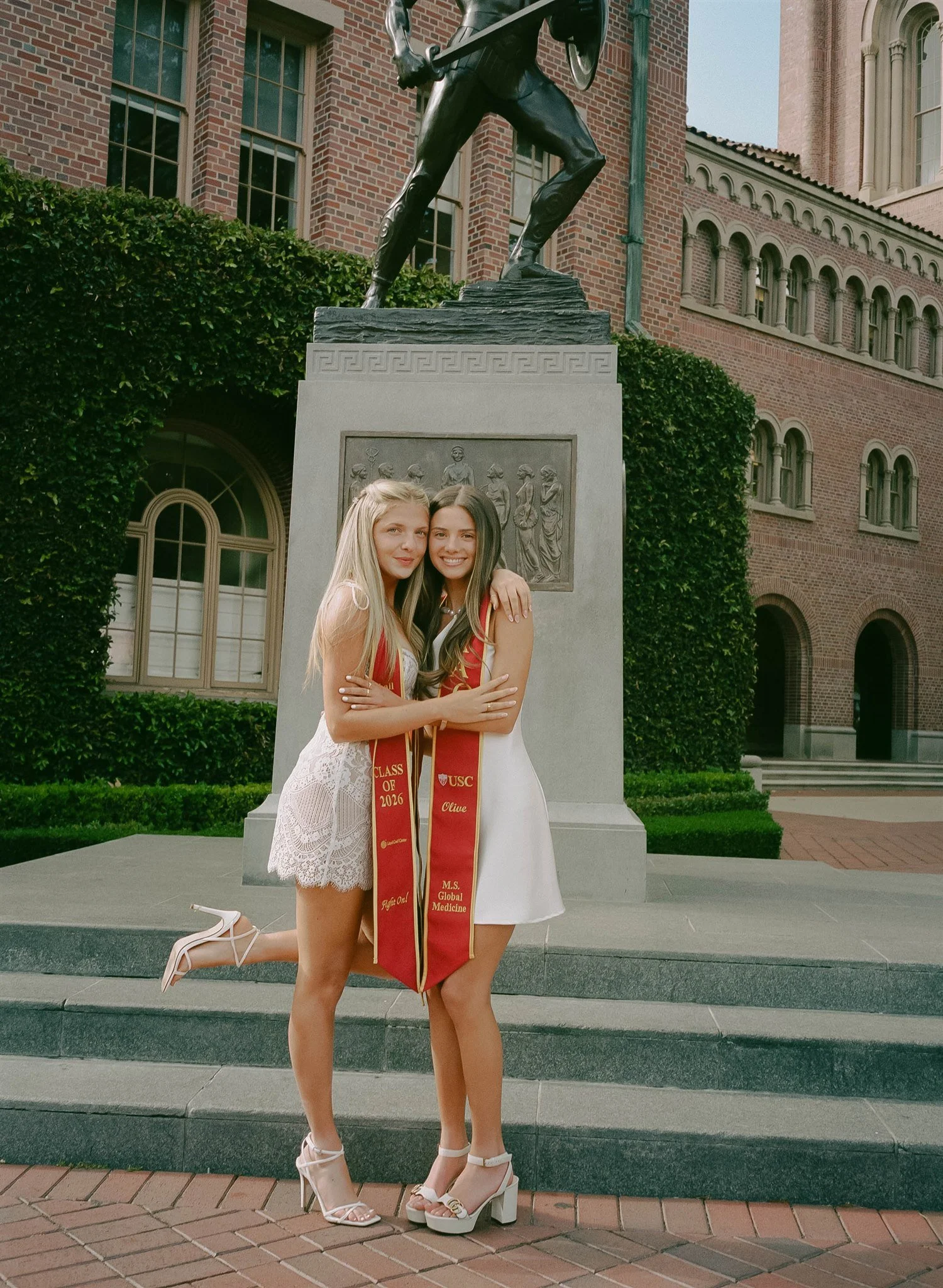 Two young women celebrating graduation outdoors, standing in front of a statue of a warrior holding a shield. They are smiling, hugging, and dressed in white dresses with red graduation sashes that read 'Class of 2026' and 'MS Global Medicine.' They 