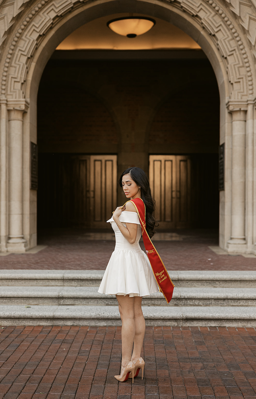 A young woman in a white dress and nude high heels standing outside a historic building, wearing a red graduation stole with gold lettering.