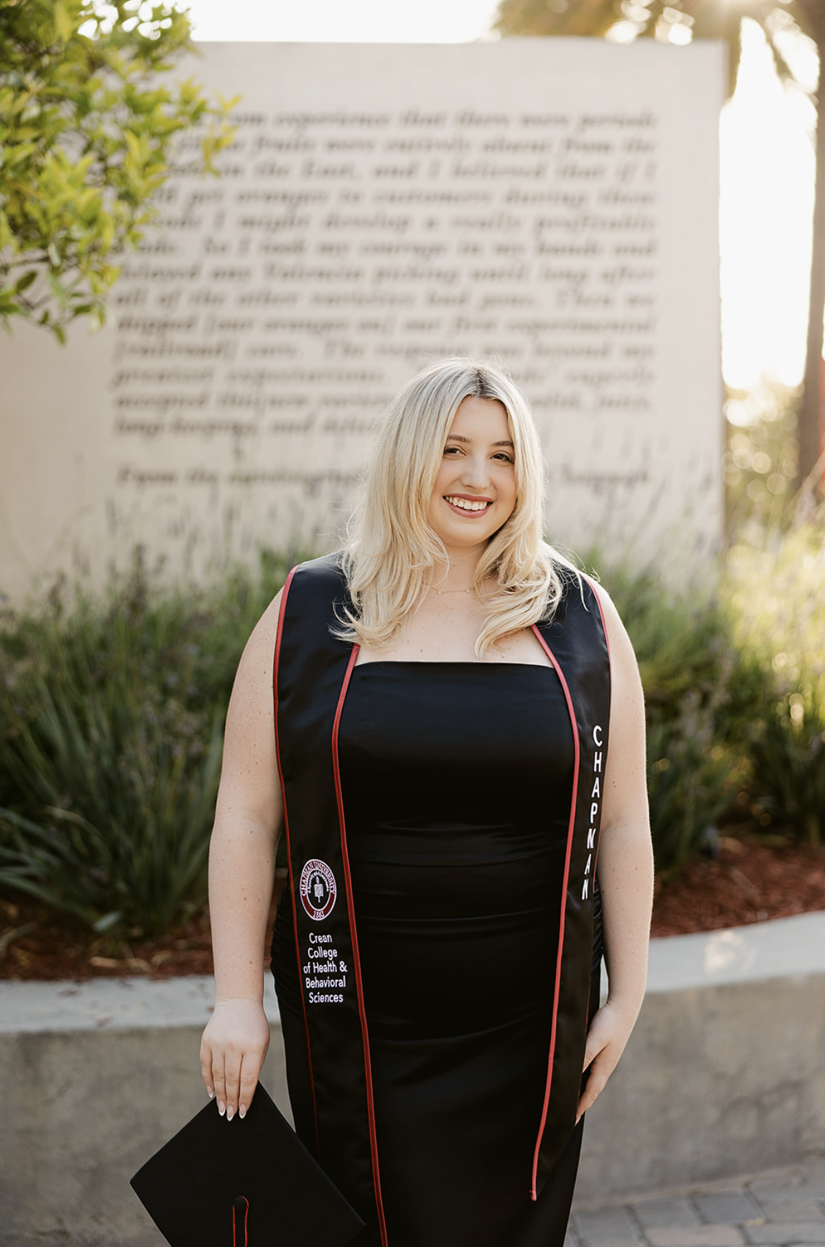 A young woman with blonde hair smiling in graduation attire, holding a cap, standing outdoors in front of a large blurred text sign and greenery.