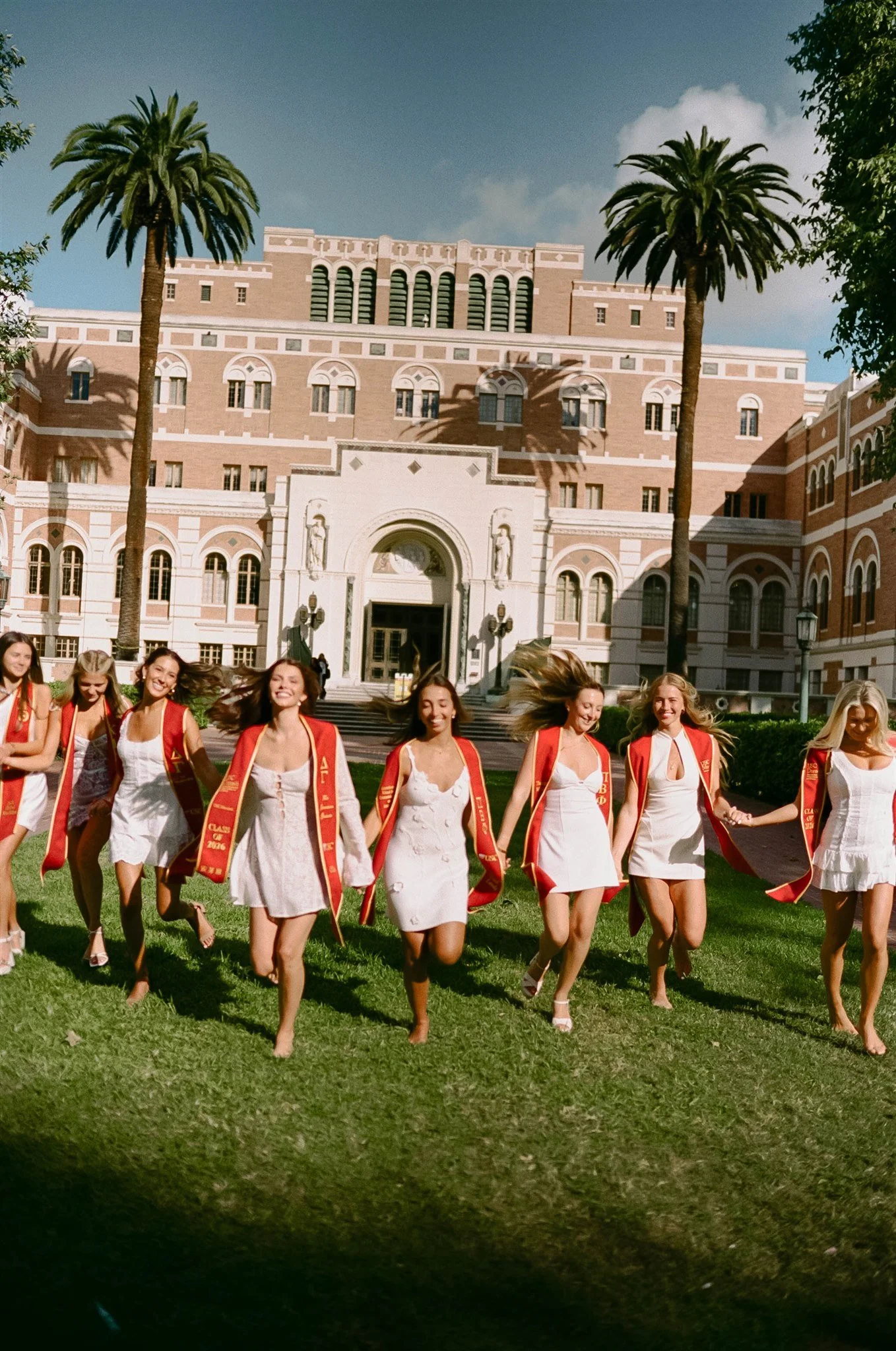 A group of women in white dresses with red sashes walking together on a green lawn in front of a large historic building with arched windows and palm trees.