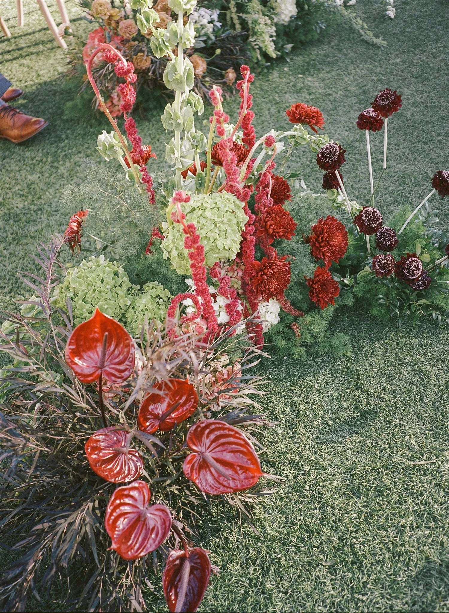 A colorful arrangement of various flowers and foliage on a grassy surface, with brown leather shoes visible in the corner.