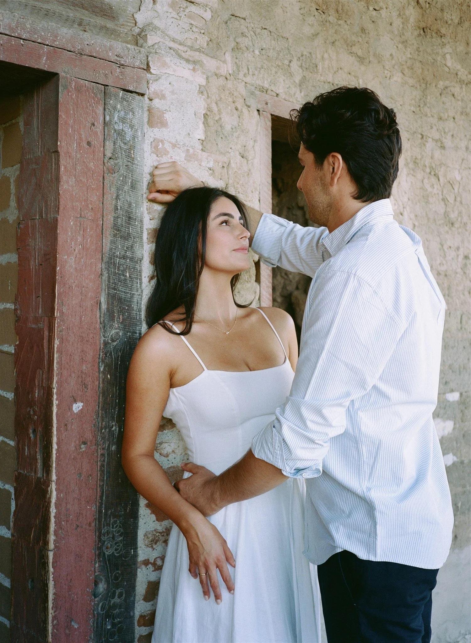 A man and woman standing close to each other against a rustic brick wall. The woman is wearing a white dress and the man a light blue striped shirt. The woman looks up at the man, who is smiling and touching her forehead gently. The scene is intimate