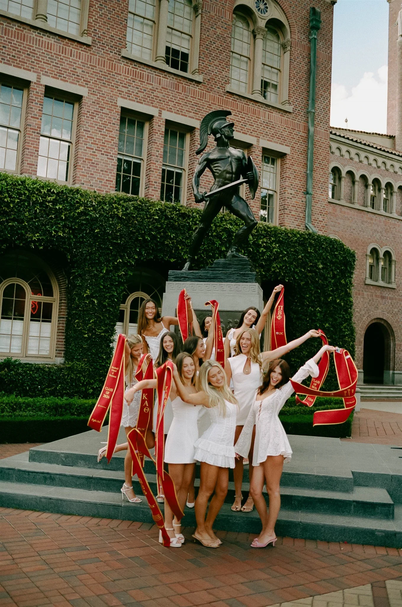 A group of young women dressed in white, celebrating outdoors in front of a large statue of a Greek warrior or soldier on a platform, with a brick building in the background.
