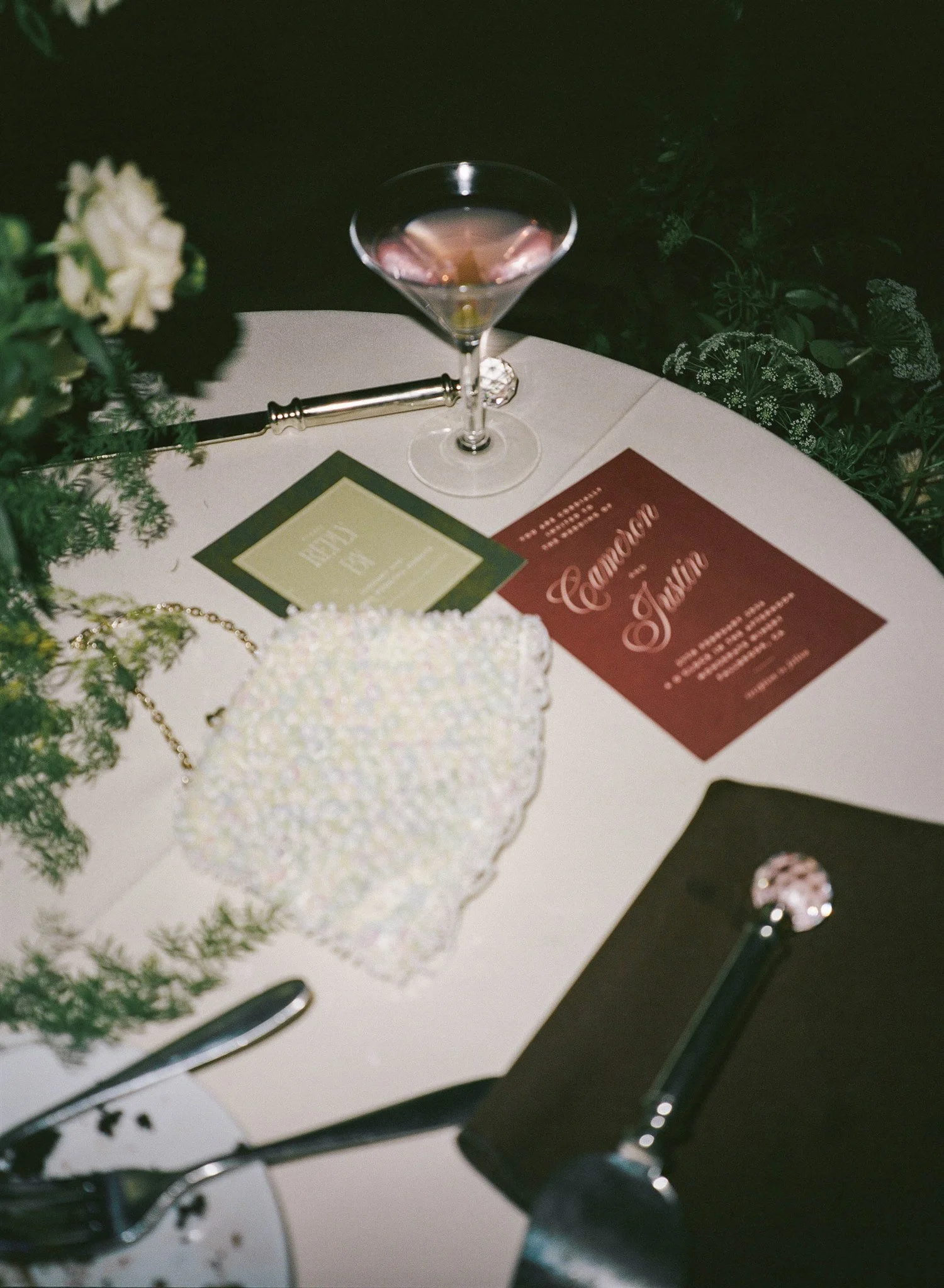 A fancy cocktail glass filled with a pink drink, on a table with flowers, menus, and jewelry, in a dimly lit setting.