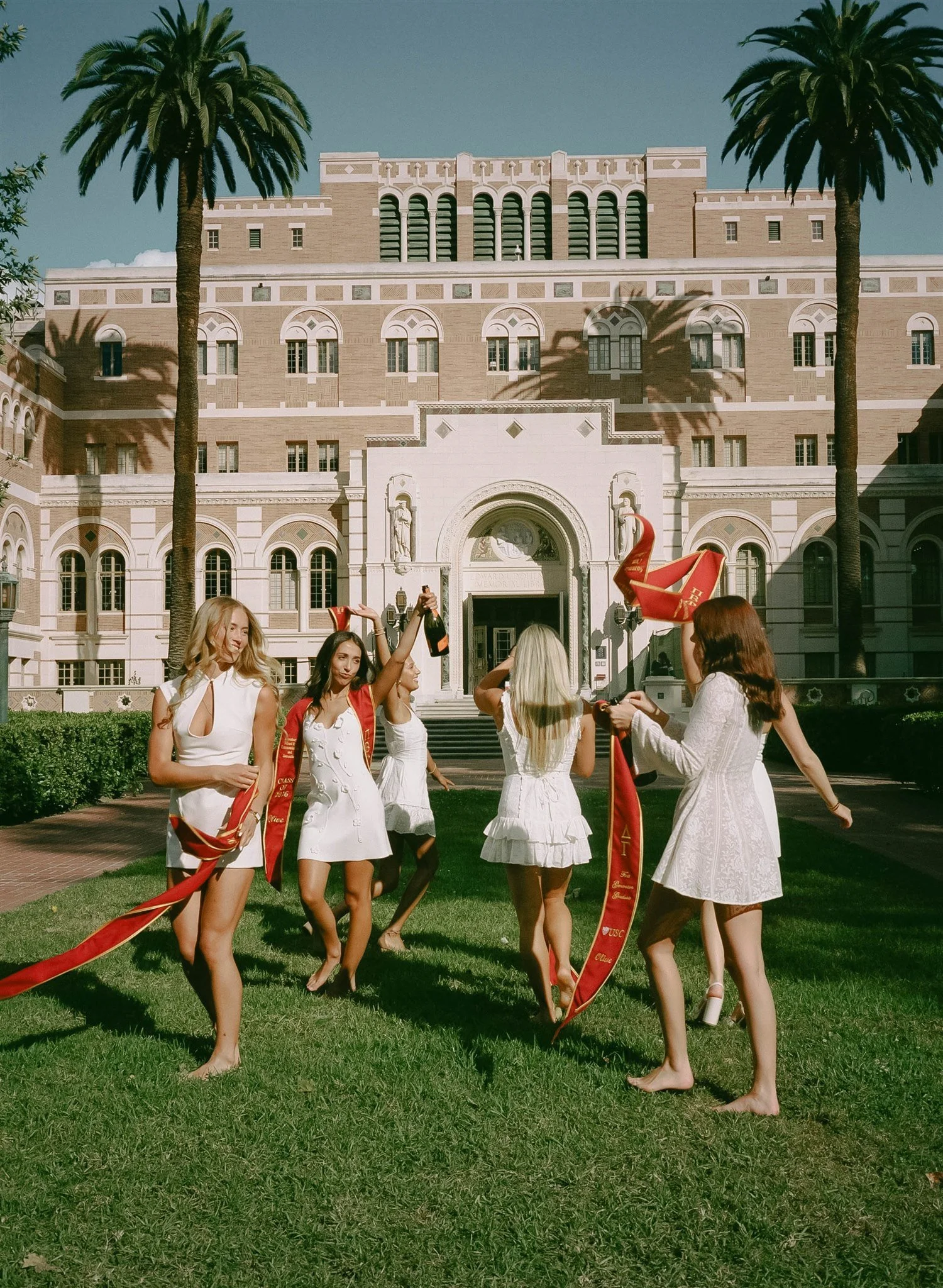 A group of women dressed in white dresses holding red sashes and champagne bottles, celebrating on the grass in front of a historic building with palm trees.