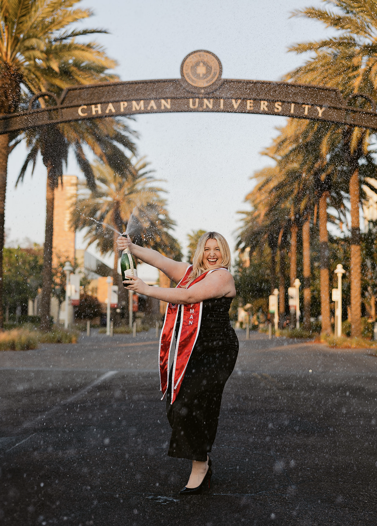 A woman celebrating graduation on a street, wearing a cap and gown with a graduation stole, holding a champagne bottle, and spraying champagne. The street is lined with palm trees and an arch overhead reads "Chapman University."