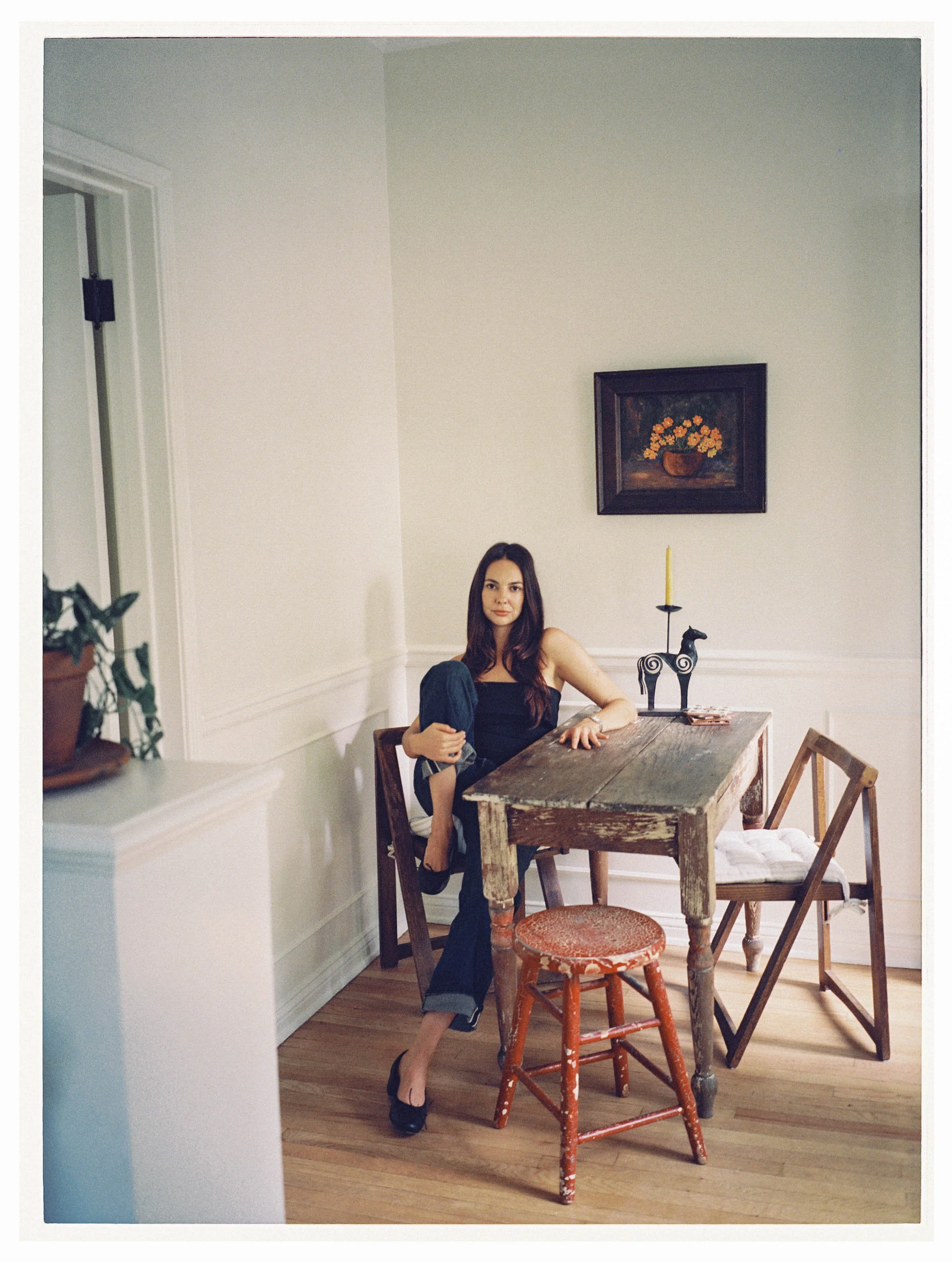 A woman with long brown hair sitting at a rustic wooden table in a room with white walls, hardwood floors, a framed painting of yellow flowers on the wall, and various chairs, including a red worn stool and a wooden chair.