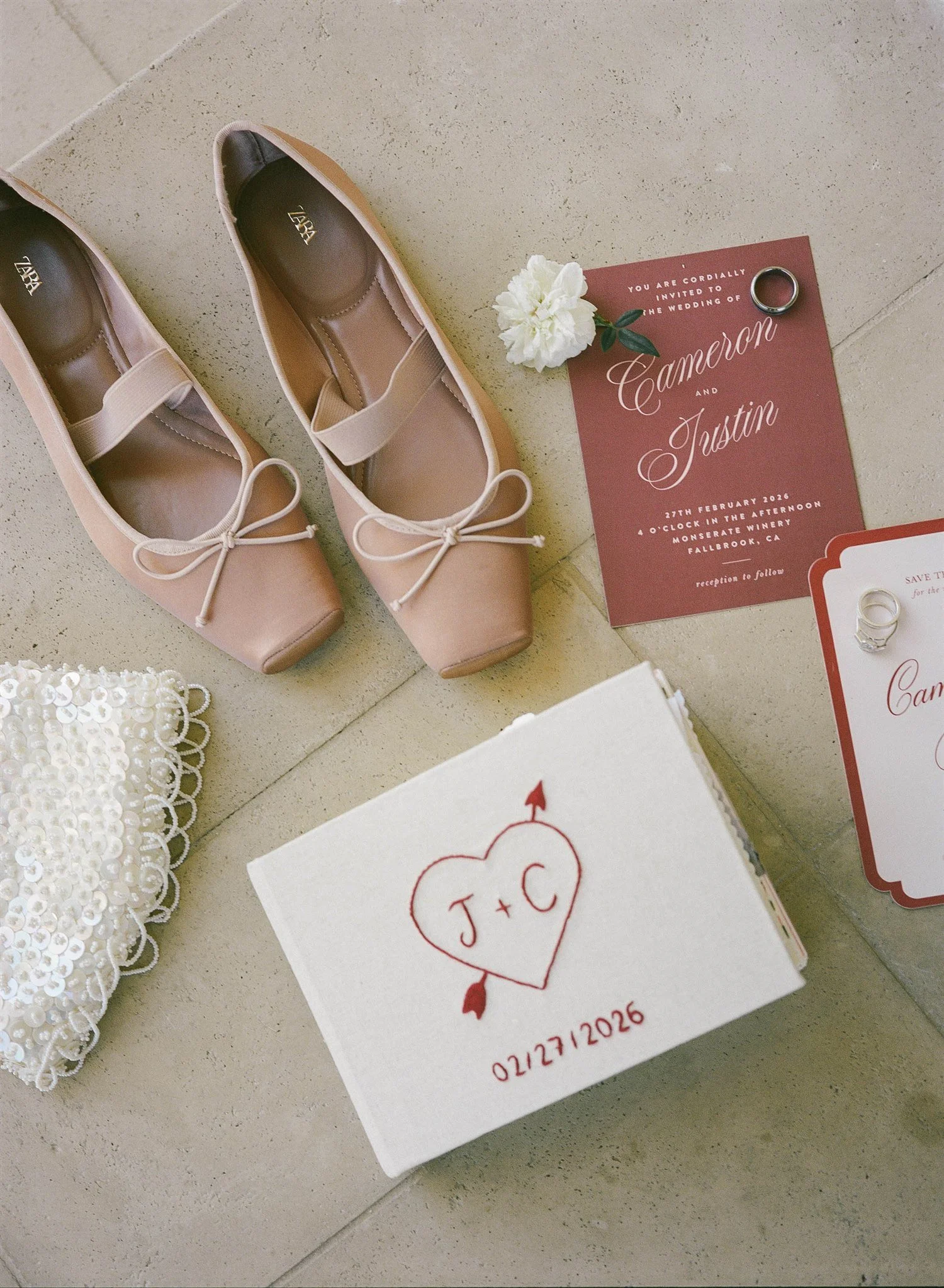 A pair of pink ballet flats with bows, a wedding invitation for Cameron and Trustin, a white flower, and a wedding ring placed on the invitation, with an additional card that has a heart with initials J and C and a date.