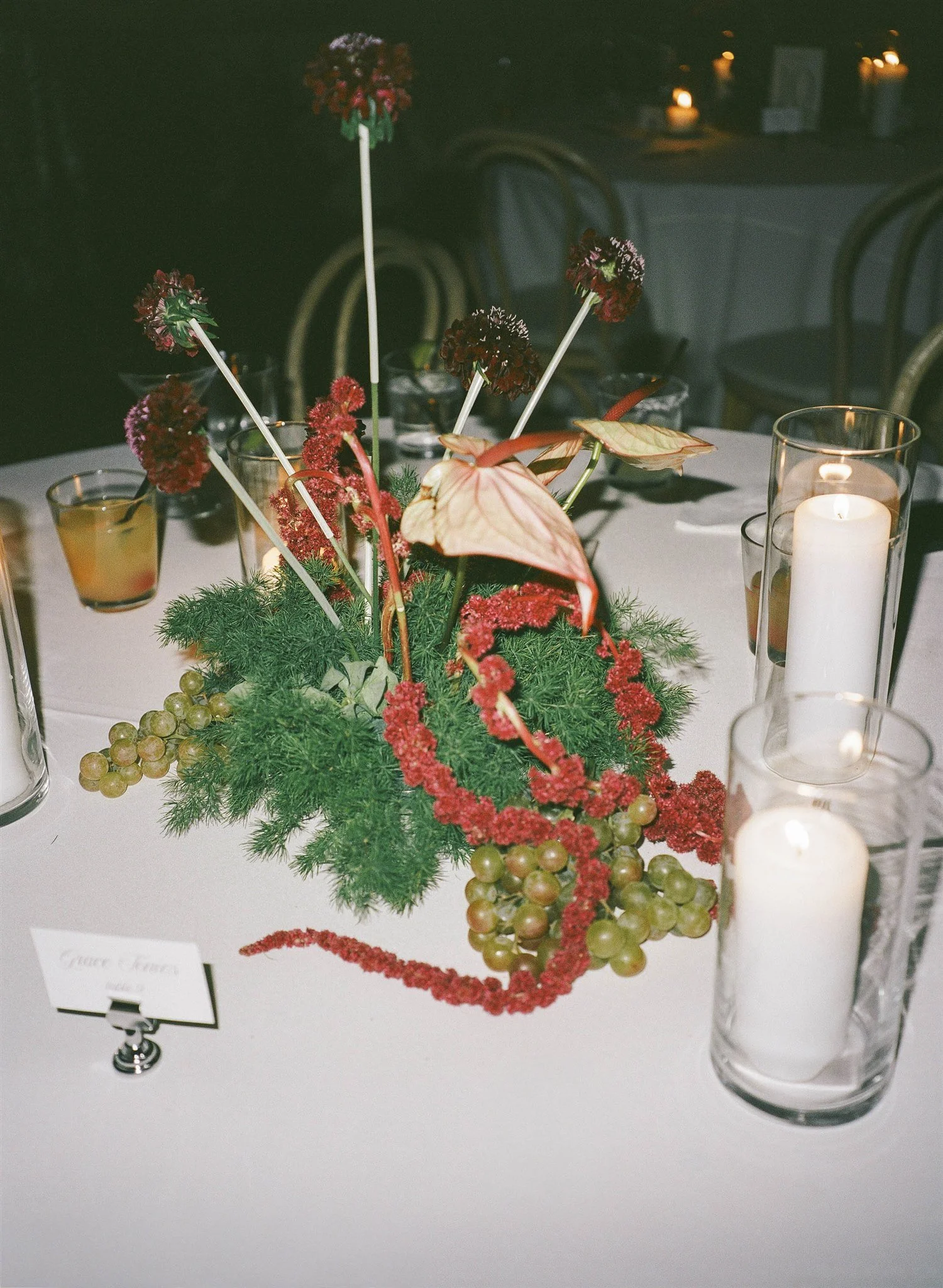 A floral centerpiece with red, burgundy, and green foliage on a white table, surrounded by candles in glass holders and small glasses with drinks, in a dimly lit setting.