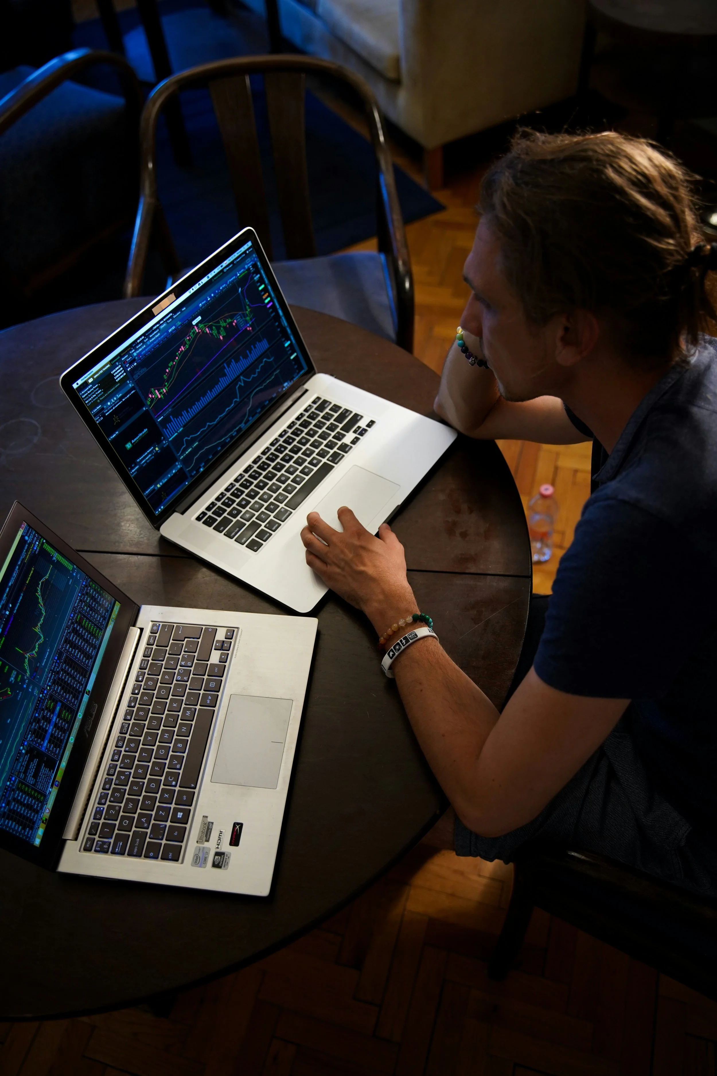 A young man with tattoos and bracelets sits at a dark wooden table, analyzing stock market charts on two open laptops. The screens display financial data and stock price graphs. The room has wooden flooring, and a bottle of water and a pink bottle cap are visible on the floor.