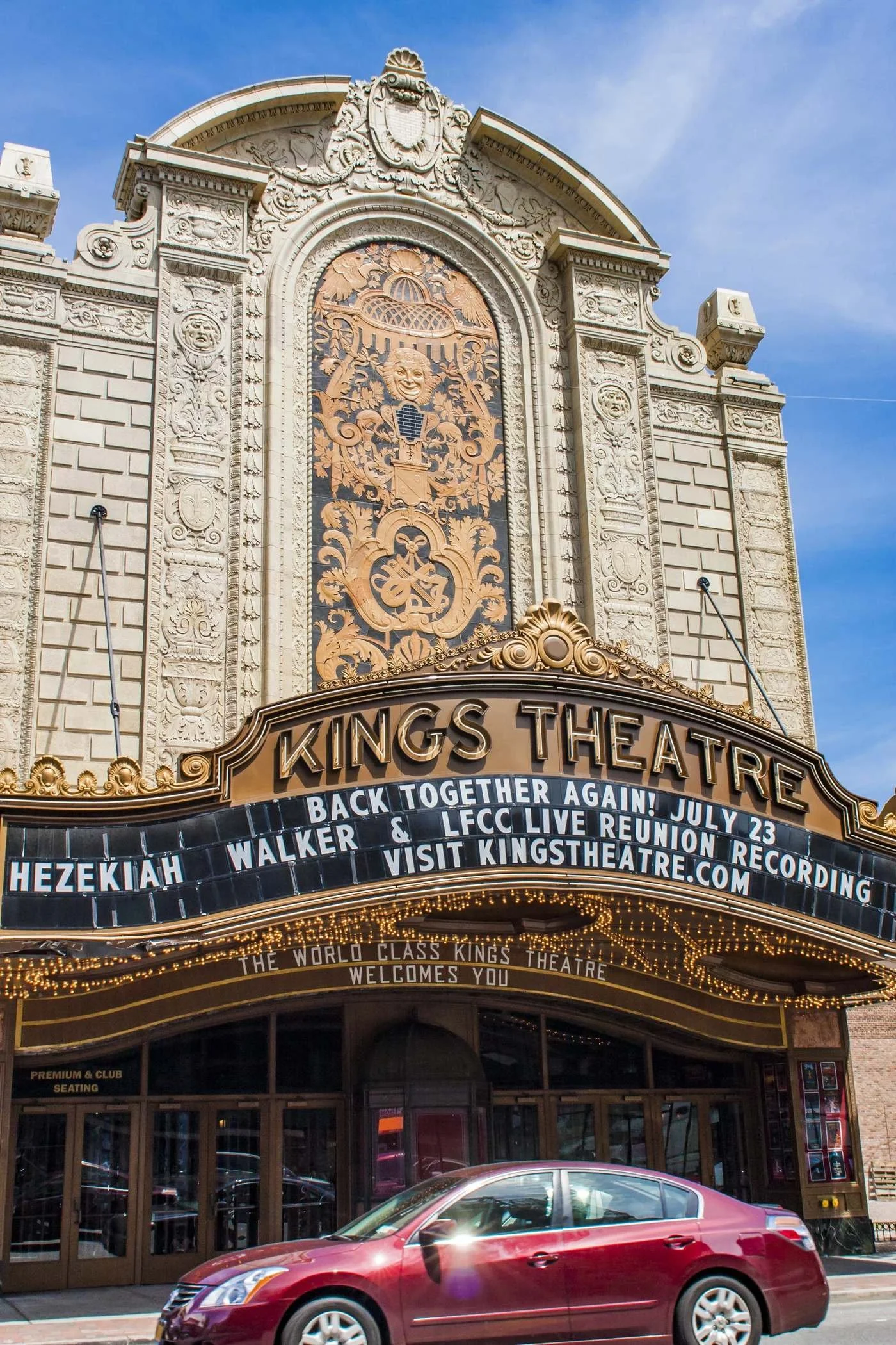 The image shows the ornate entrance of Kings Theatre with decorative carvings and a marquee displaying upcoming show information. A red car is parked in front of the theater.