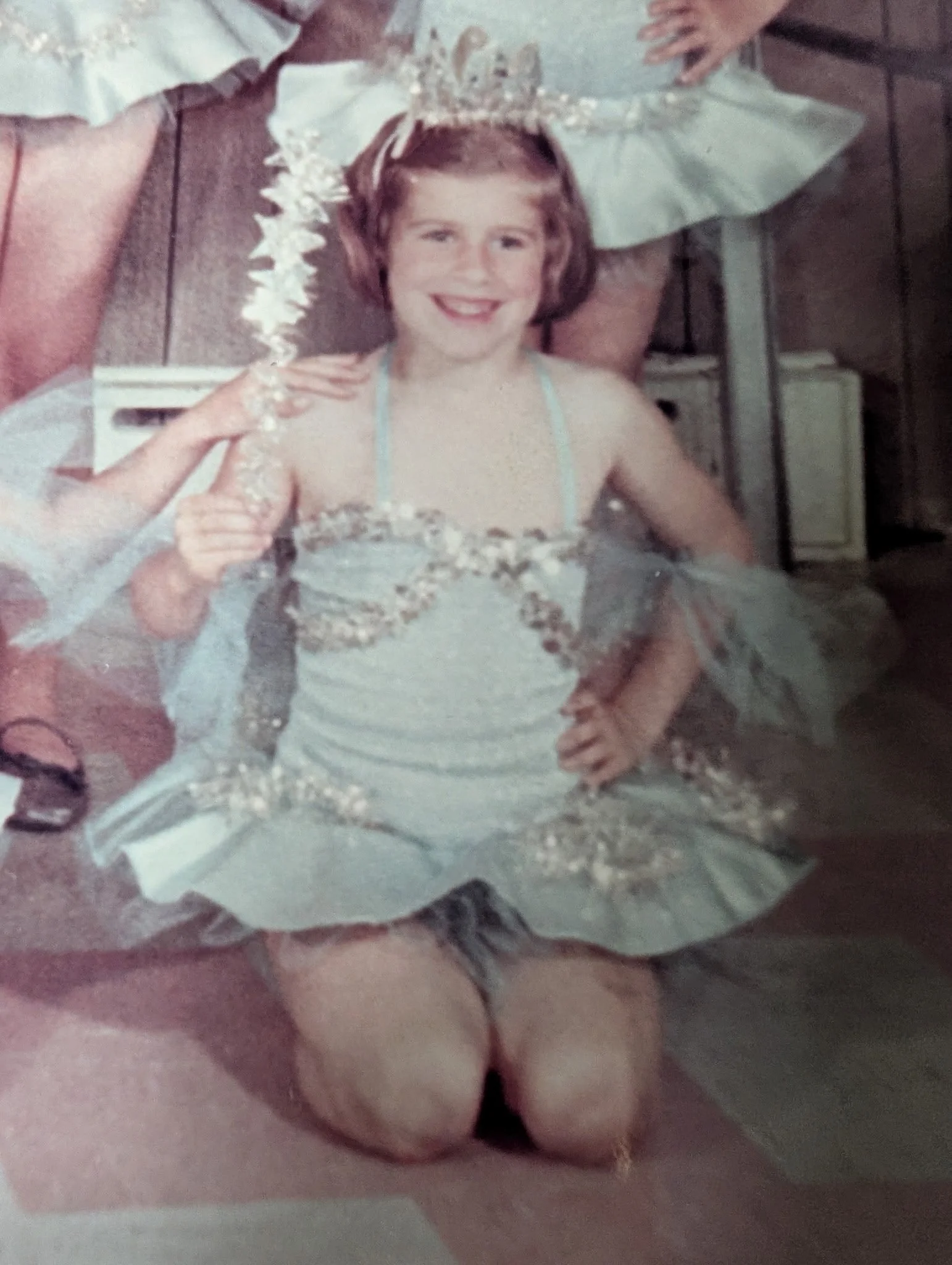 A young girl dressed in a ballet costume with a tiara on her head, kneeling with her hands on her hips, smiling. Behind her, an adult person is partially visible, holding her by the shoulders. The setting appears to be indoors with wood-paneled walls.