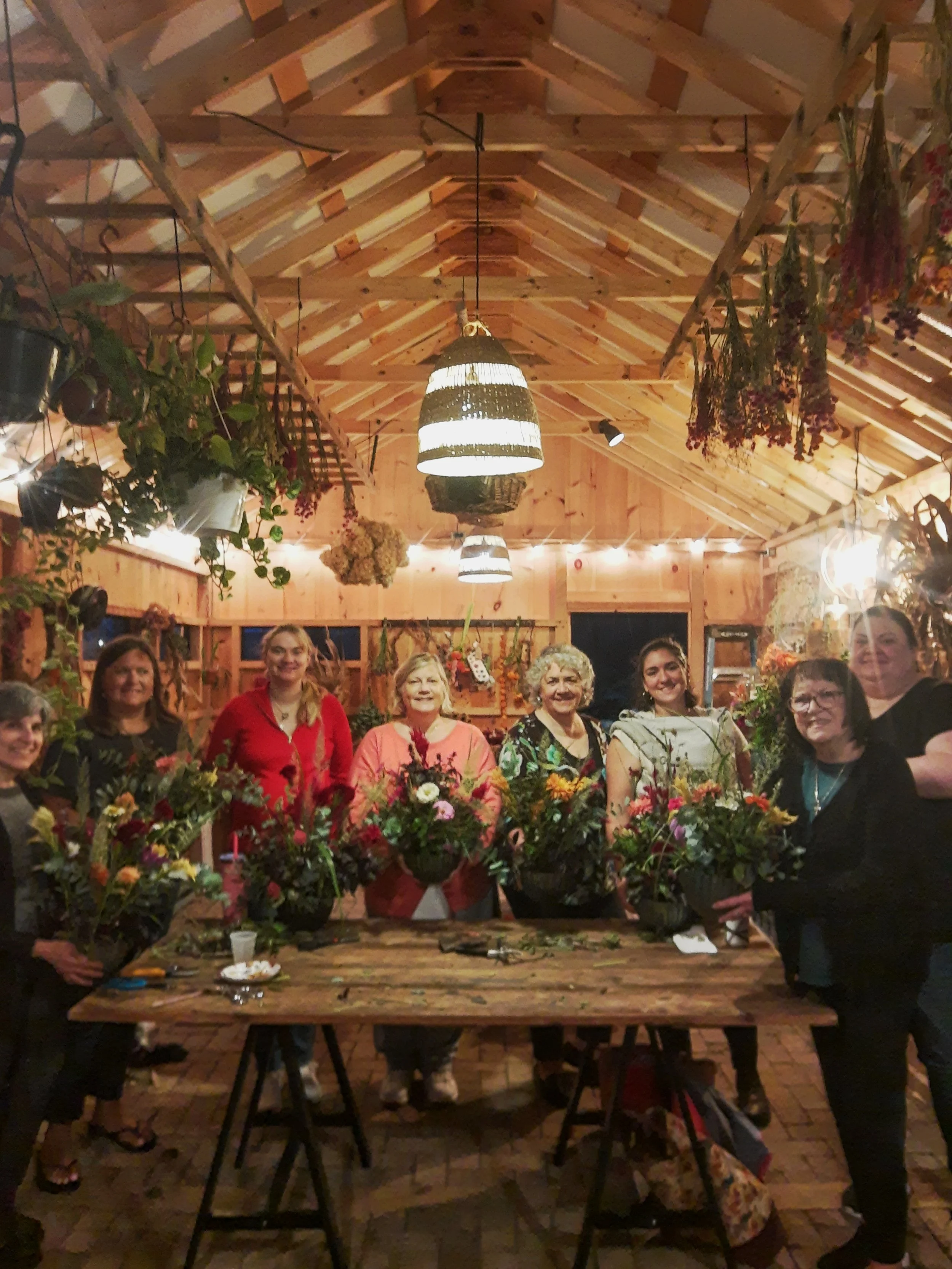 A group of women standing around a table with floral arrangements inside a warmly lit wooden greenhouse or workshop.